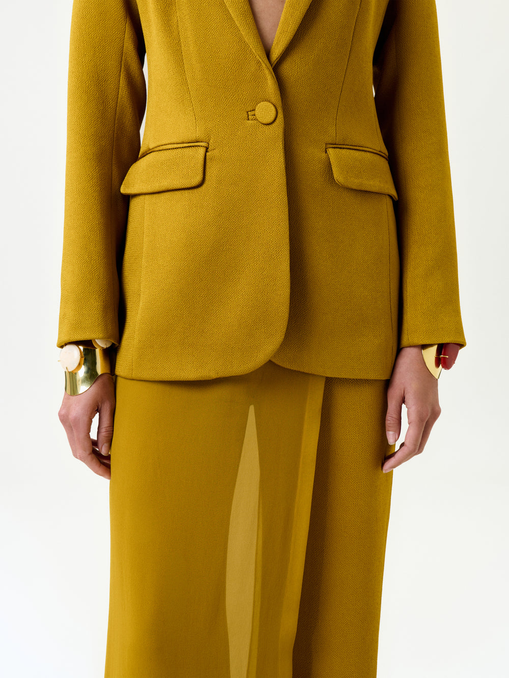 A model wears the Rebeca Jacket in mustard yellow Italian fabric with a matching skirt and gold statement bracelets, shown from shoulders to knees against a plain white background.