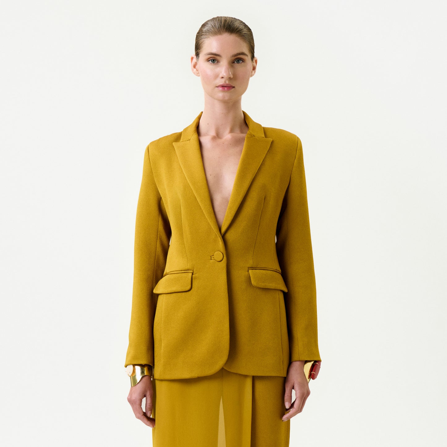 A person stands facing forward in the Rebeca Jacket—a mustard yellow Italian fabric jacket with matching wide-leg trousers and chunky bracelets. Set against a plain white background, this tailored outfit delivers a modern, true-to-size fit.