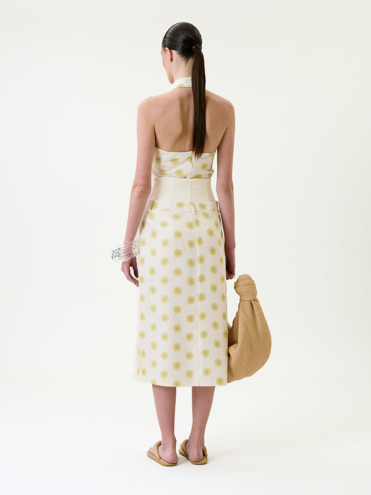 A woman stands facing away, wearing the Lucena Skirt—a white, mid-length piece with yellow circular patterns—paired with a matching halter-neck top, tan sandals, a white bracelet, and carrying a textured tan bag. Her hair is styled in a low ponytail.