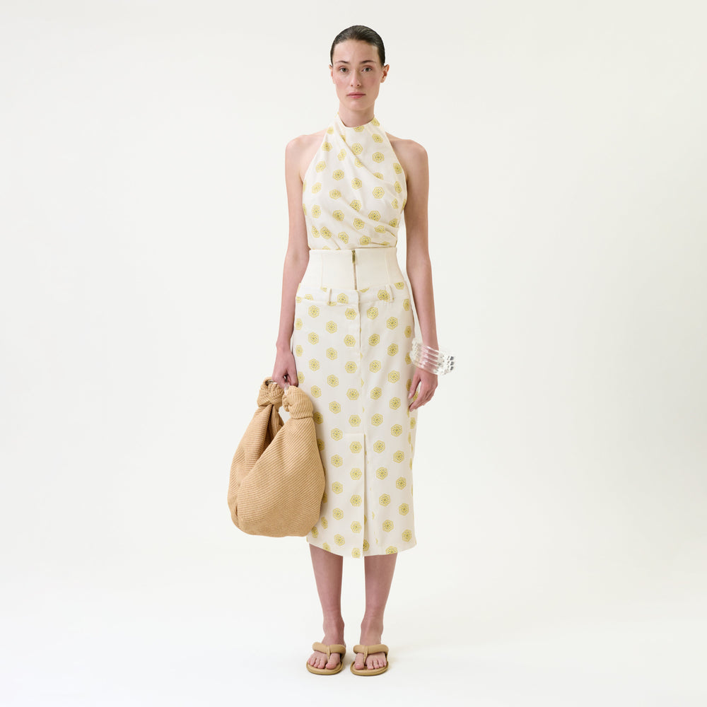 A woman stands against a plain white background, wearing the Lucena Skirt—a sleeveless halter top and matching midi skirt set with yellow circular patterns. She holds a large tan textured bag and accessorizes with sandals and chunky white bracelets.