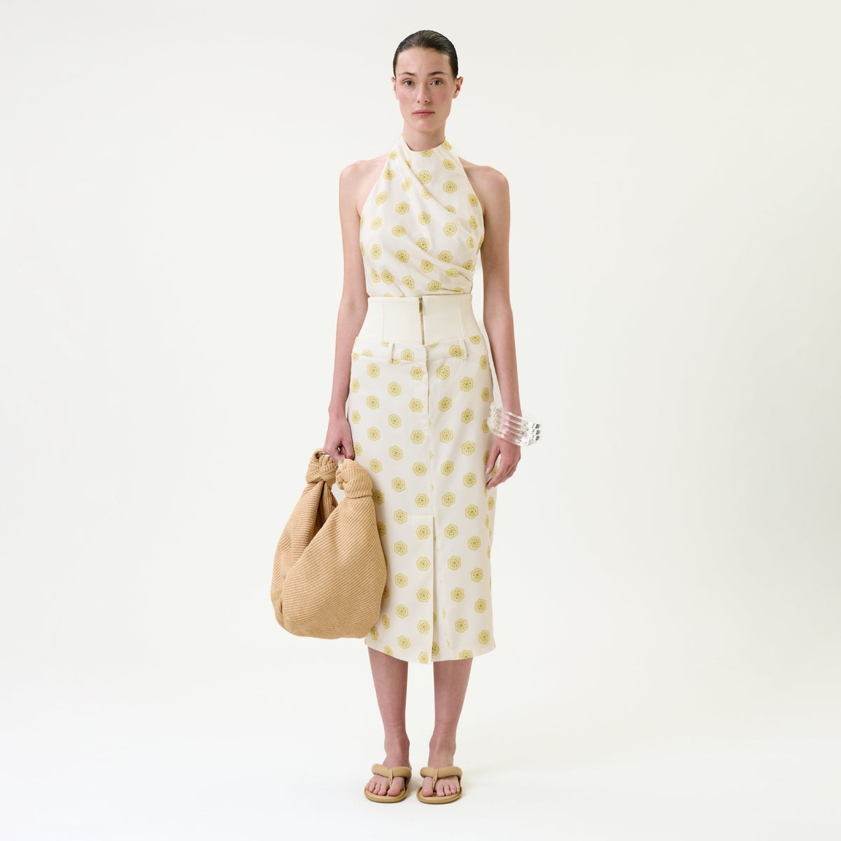 A woman stands against a plain white background, wearing the Lucena Skirt—a sleeveless halter top and matching midi skirt set with yellow circular patterns. She holds a large tan textured bag and accessorizes with sandals and chunky white bracelets.