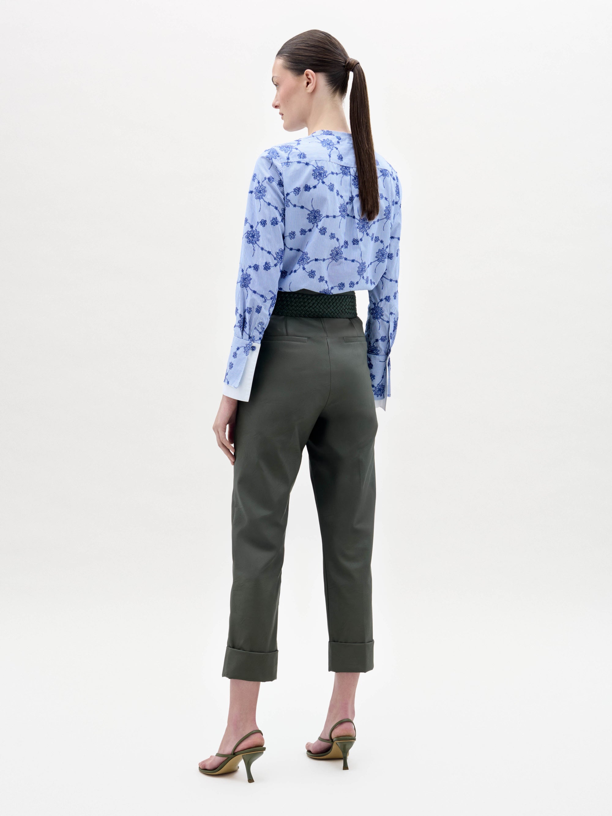 A woman stands against a plain background wearing a blue floral blouse, the Moad Pant—dark green, high-waisted with a wide belt featuring a gold plate and fringe—and heeled sandals.