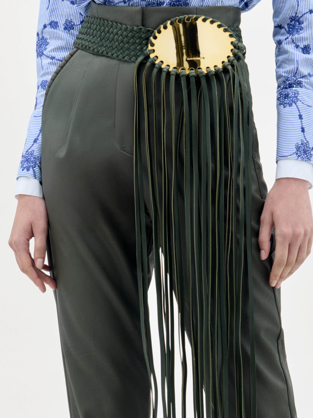 A woman stands against a plain background wearing a blue floral blouse, the Moad Pant—dark green, high-waisted with a wide belt featuring a gold plate and fringe—and heeled sandals.