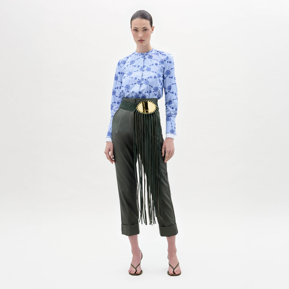A woman stands against a plain background wearing a blue floral blouse, the Moad Pant—dark green, high-waisted with a wide belt featuring a gold plate and fringe—and heeled sandals.