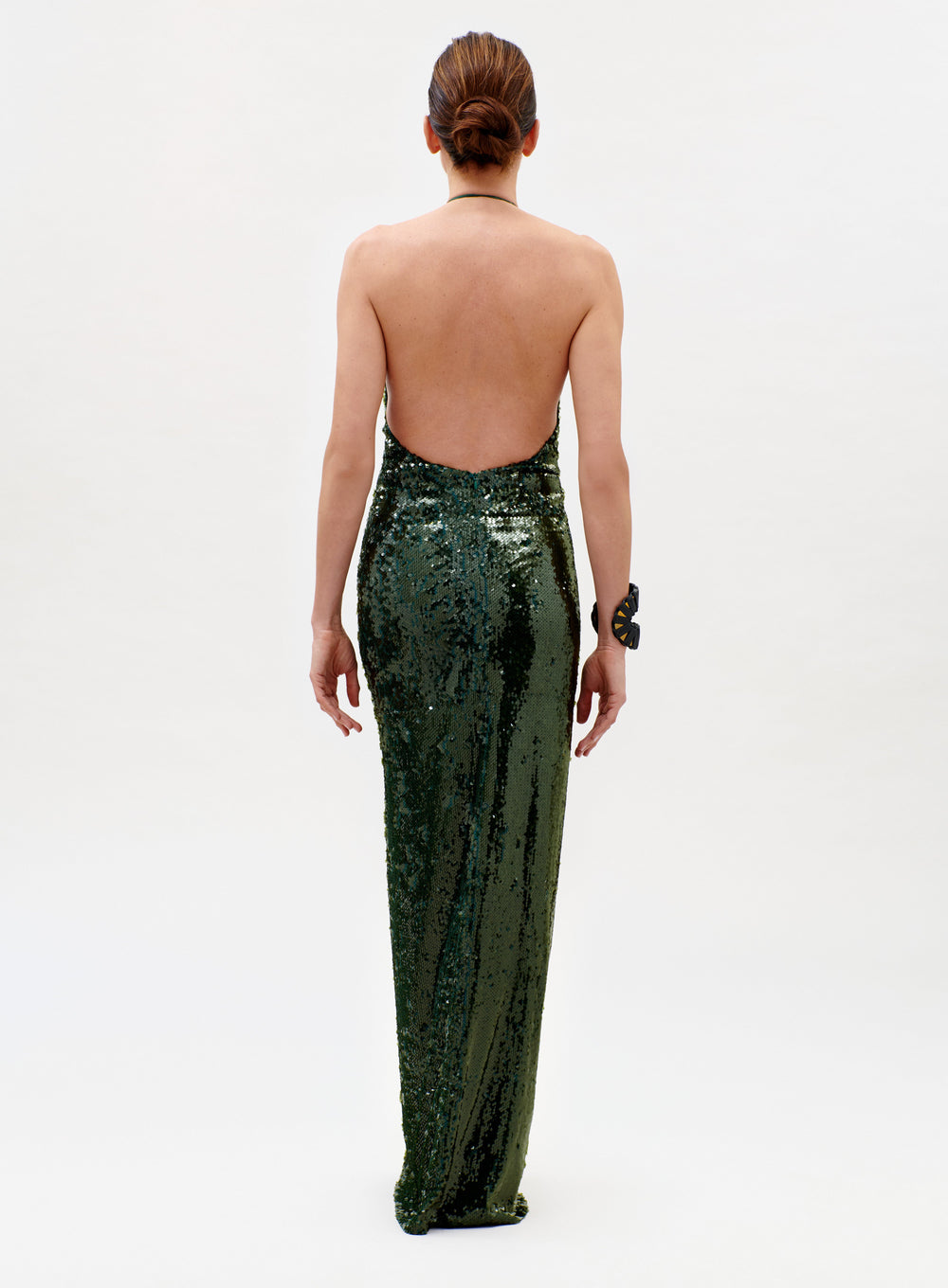 A woman faces away in the Rowan Dress, a sparkling dark green backless gown. Her hair is styled in a low bun, and she wears a large bracelet on her left wrist against a plain white background.