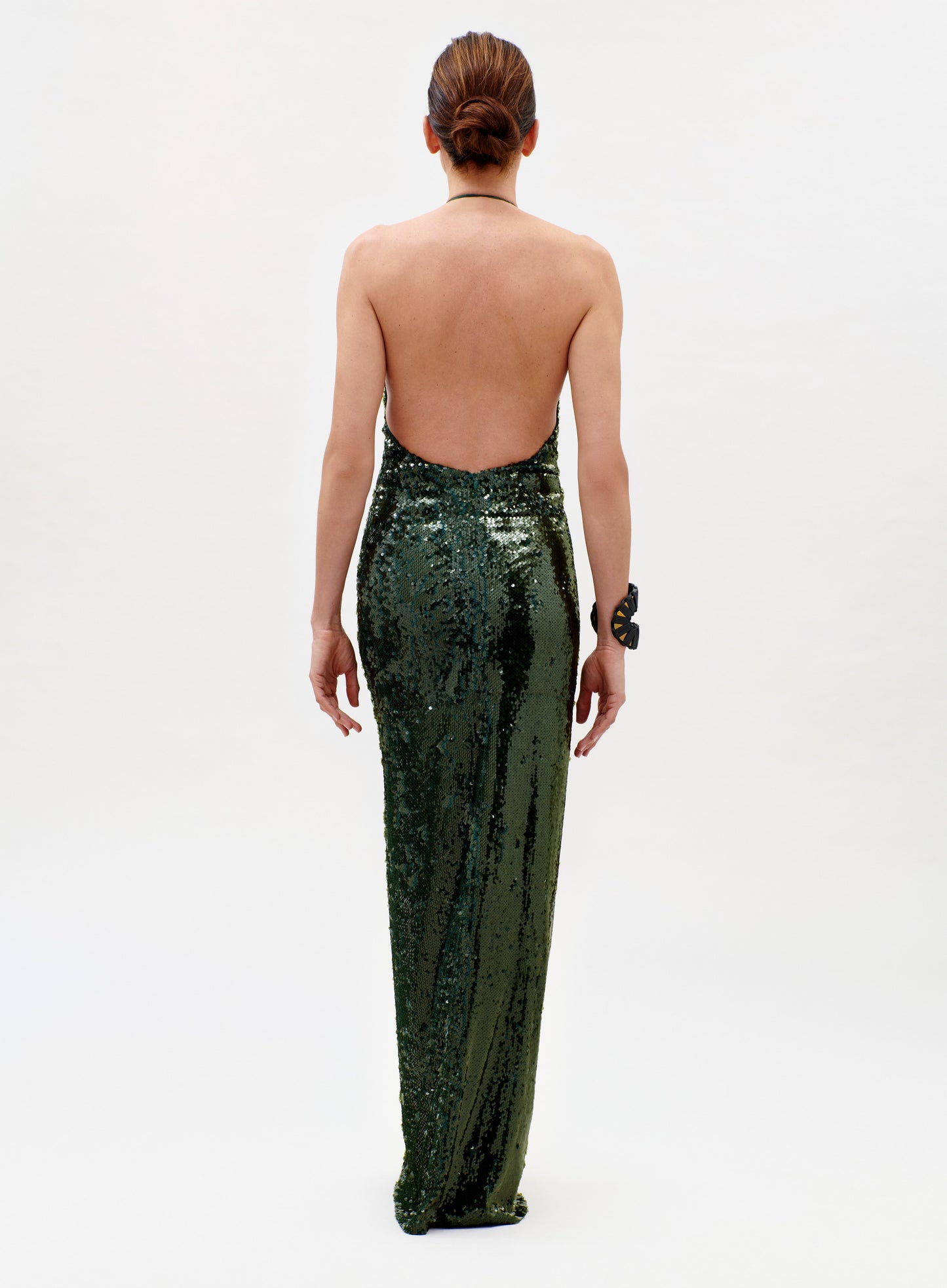 A woman faces away in the Rowan Dress, a sparkling dark green backless gown. Her hair is styled in a low bun, and she wears a large bracelet on her left wrist against a plain white background.