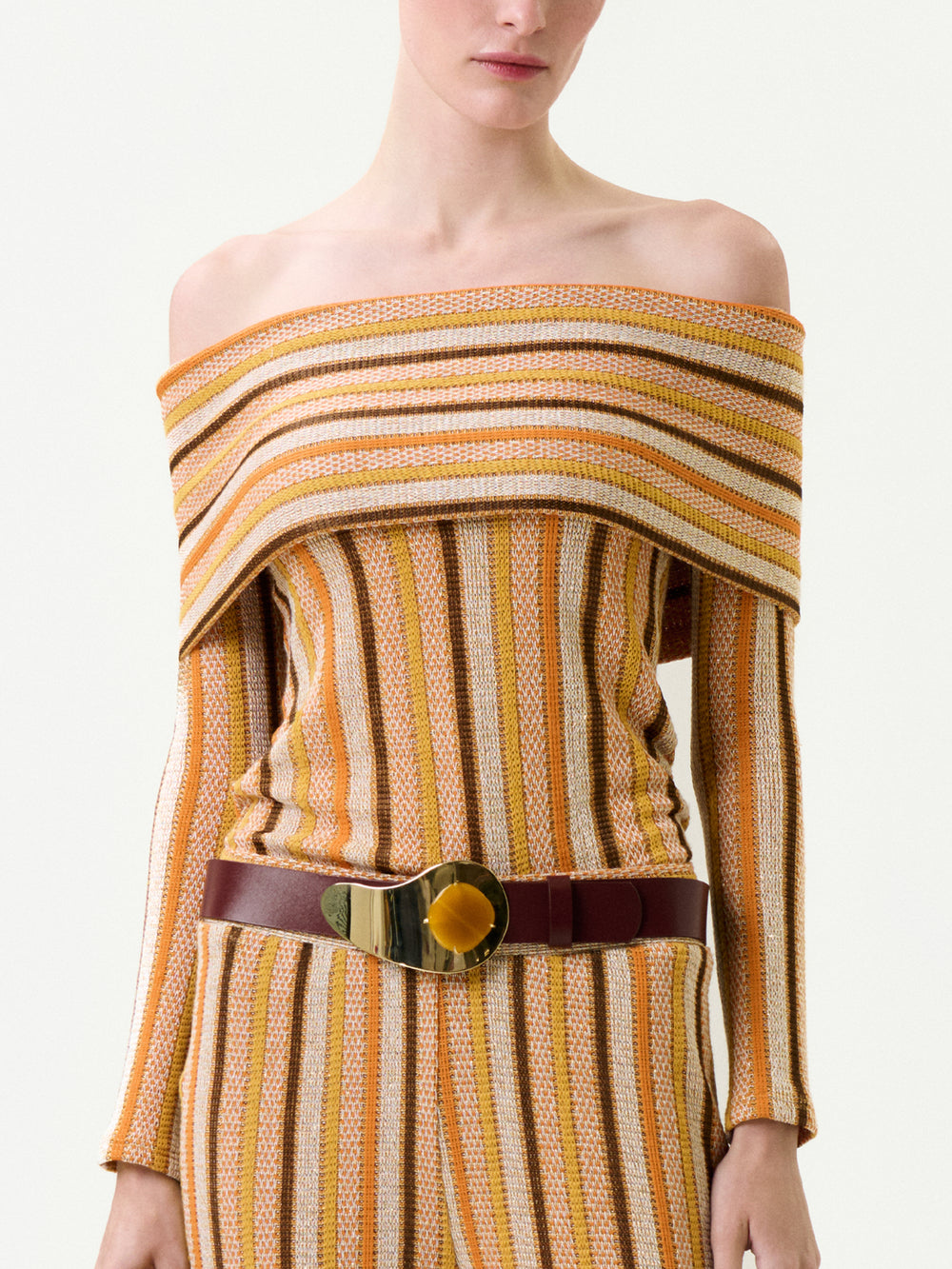 A woman wears the Maitenea Top—an off-the-shoulder, long-sleeved knit with vertical yellow and brown stripes—paired with matching pants and a wide brown belt with a gold buckle. Made in Colombia with a true-to-size fit.