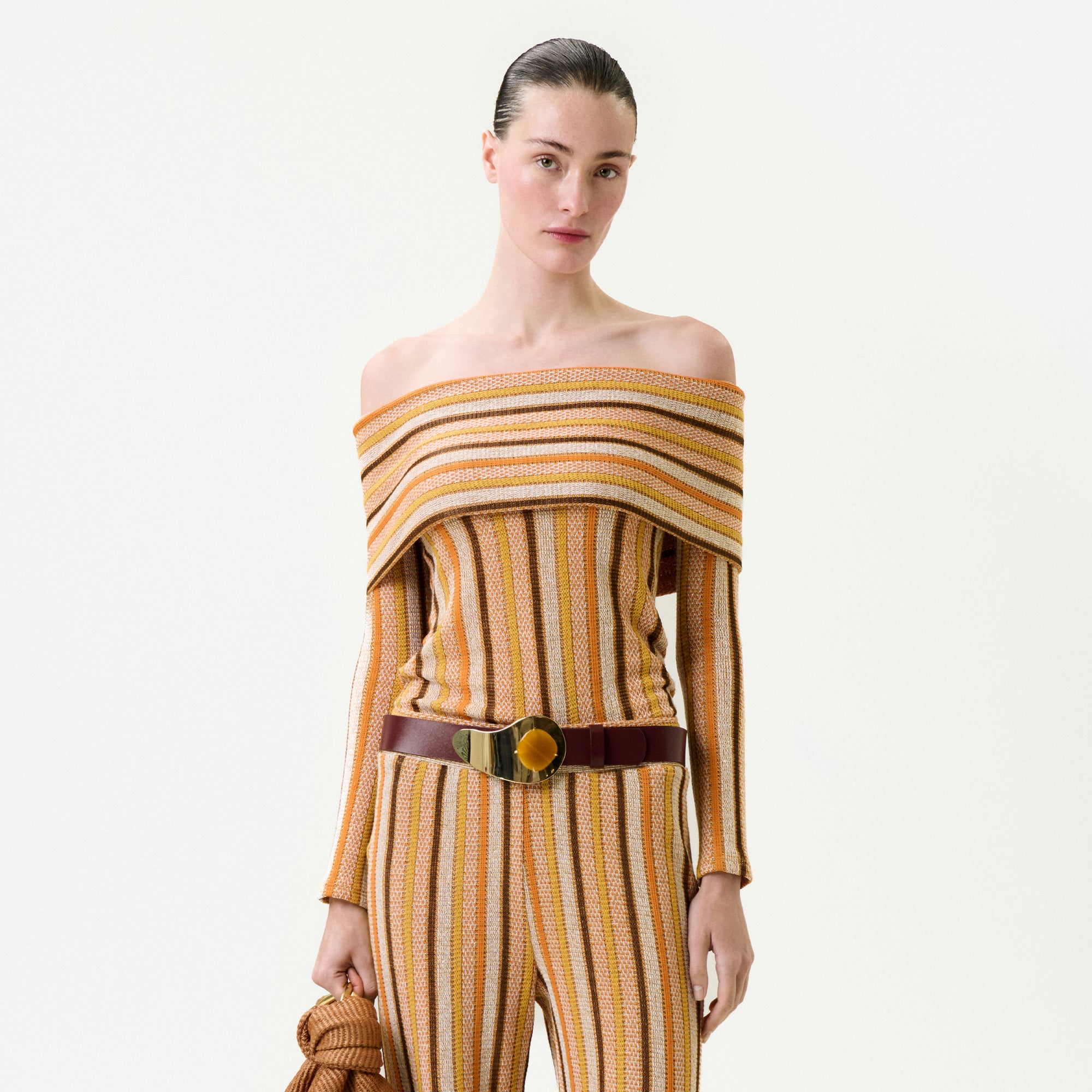 Wearing the Maitenea Top—a striped off-the-shoulder design in orange, yellow, and beige made from premium Italian fabric with a true-to-size fit—paired with matching pants, a wide brown belt, and a textured bag.