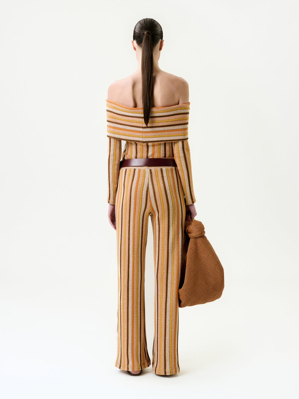 A person with long, straight brown hair in a ponytail is seen from behind, wearing the Naoki Pant—a matching flared style and off-the-shoulder striped top made of Italian fabric—while holding a large textured brown bag.