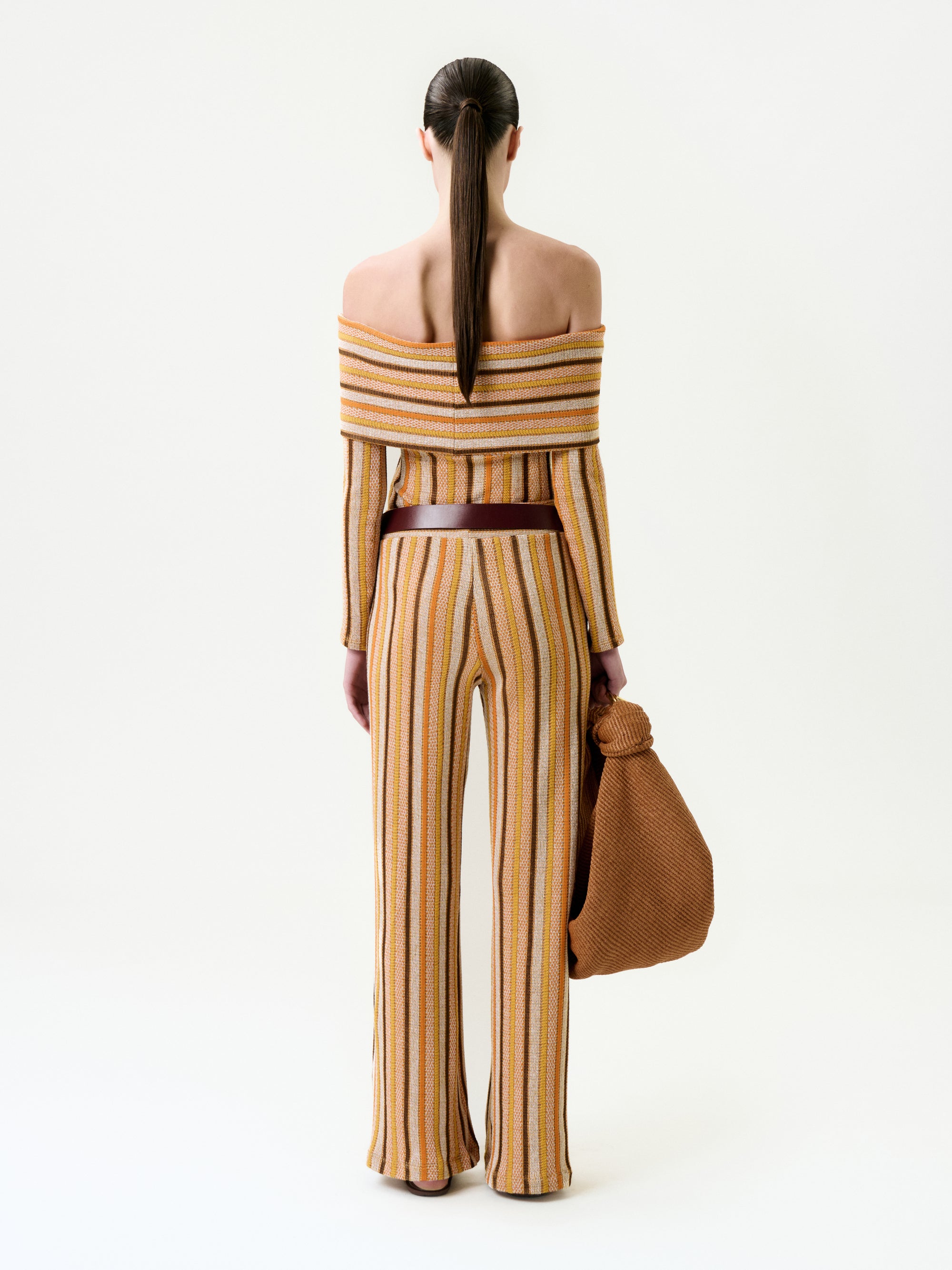 A person with long, straight brown hair in a ponytail is seen from behind, wearing the Naoki Pant—a matching flared style and off-the-shoulder striped top made of Italian fabric—while holding a large textured brown bag.