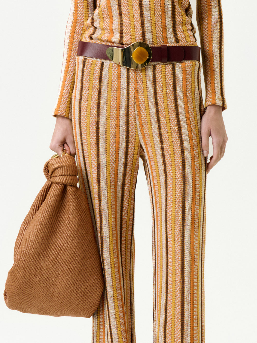 Wearing the Naoki Pant made from Italian fabric in yellow, brown, and white stripes, styled with a wide brown belt and textured brown bag with a knotted handle. The photo shows only the torso and legs.