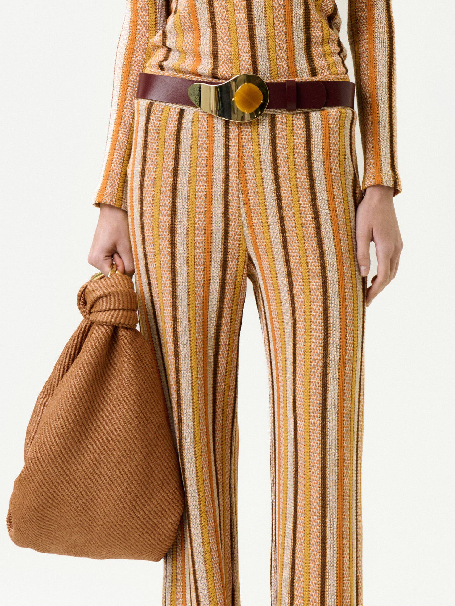 Wearing the Naoki Pant made from Italian fabric in yellow, brown, and white stripes, styled with a wide brown belt and textured brown bag with a knotted handle. The photo shows only the torso and legs.