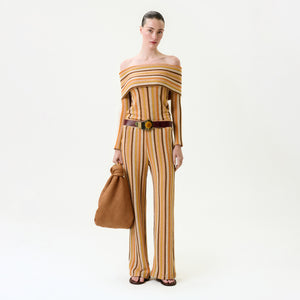 A woman stands against a white background wearing the Naoki Pant, an off-the-shoulder striped knit jumpsuit in yellow, orange, and beige. She accessorizes with a large brown textured bag, wide brown belt, and sandals. Fits true to size.