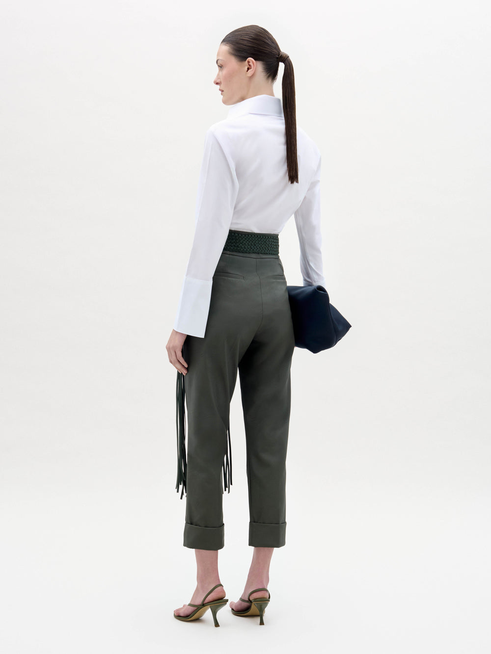 A woman stands against a white background wearing the Andrea Blouse, dark green belted pants with fringe detail, heeled sandals, and holding a dark clutch.