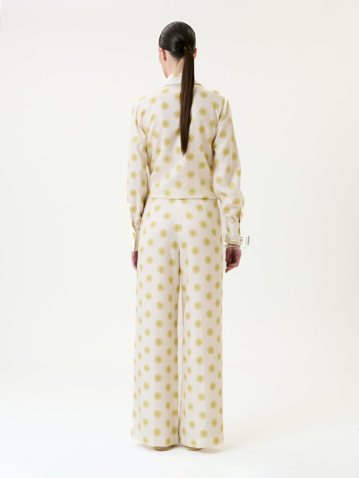A person stands facing away, wearing the Caserta Blouse—a cream-colored top with yellow circular patterns and long sleeves—paired with matching wide-legged pants. Their hair is tied back in a low ponytail, set against a plain white background.