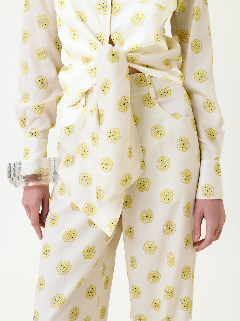 Model wearing the Caserta Blouse—a white top with a yellow geometric print, tied at the waist. Clear chunky bracelets adorn their wrist, and only the lower torso and upper legs are visible.