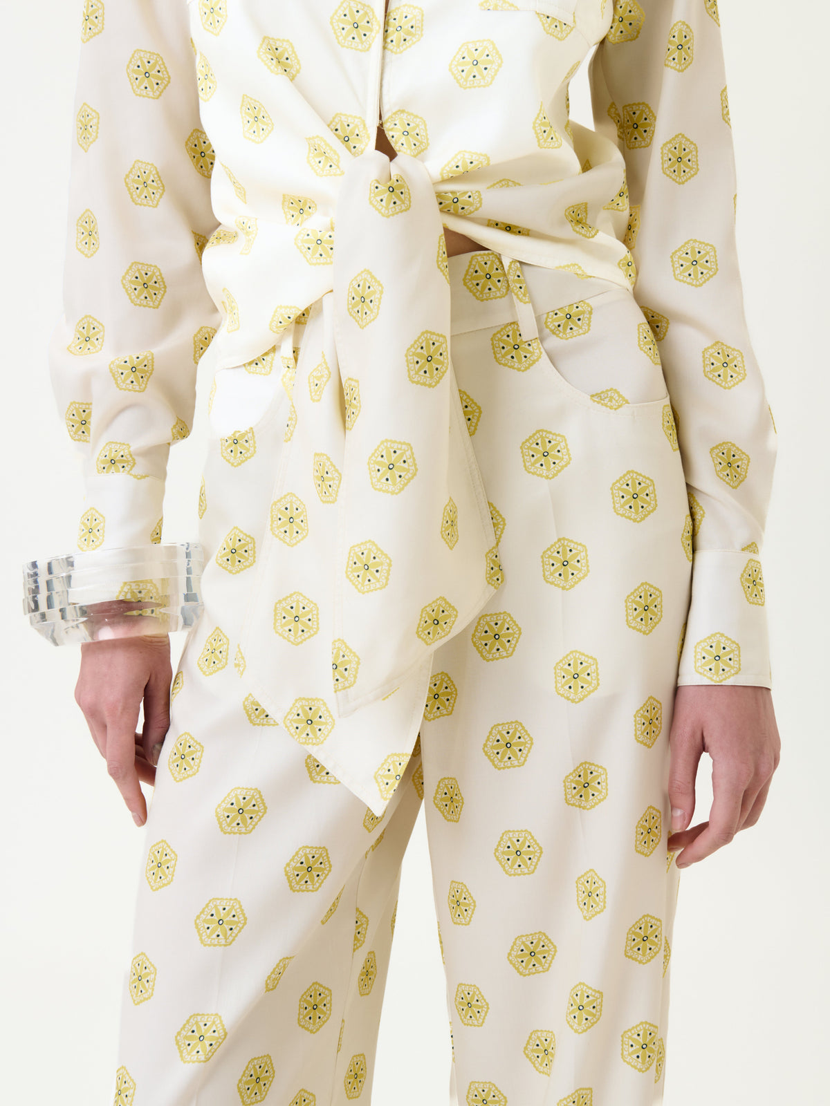 Model wearing the Caserta Blouse—a white top with a yellow geometric print, tied at the waist. Clear chunky bracelets adorn their wrist, and only the lower torso and upper legs are visible.