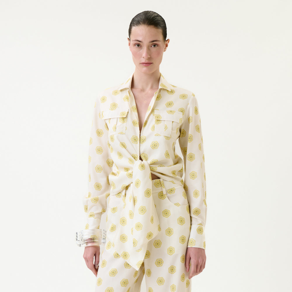 A woman with slicked-back hair wears the Caserta Blouse—a cream-colored, collared shirt with yellow circular patterns, tied at the waist—paired with matching pants, standing against a plain white background.