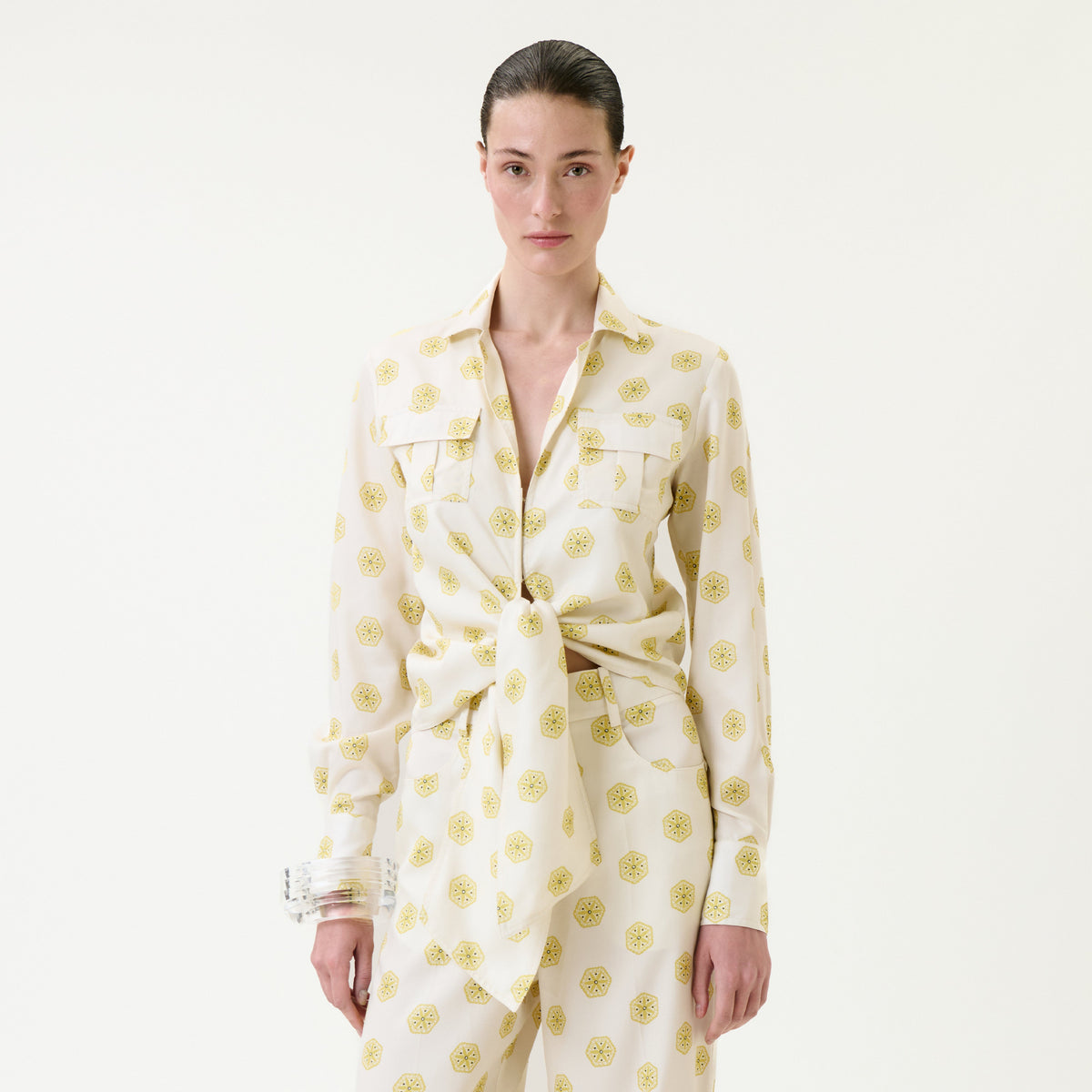 A woman with slicked-back hair wears the Caserta Blouse—a cream-colored, collared shirt with yellow circular patterns, tied at the waist—paired with matching pants, standing against a plain white background.