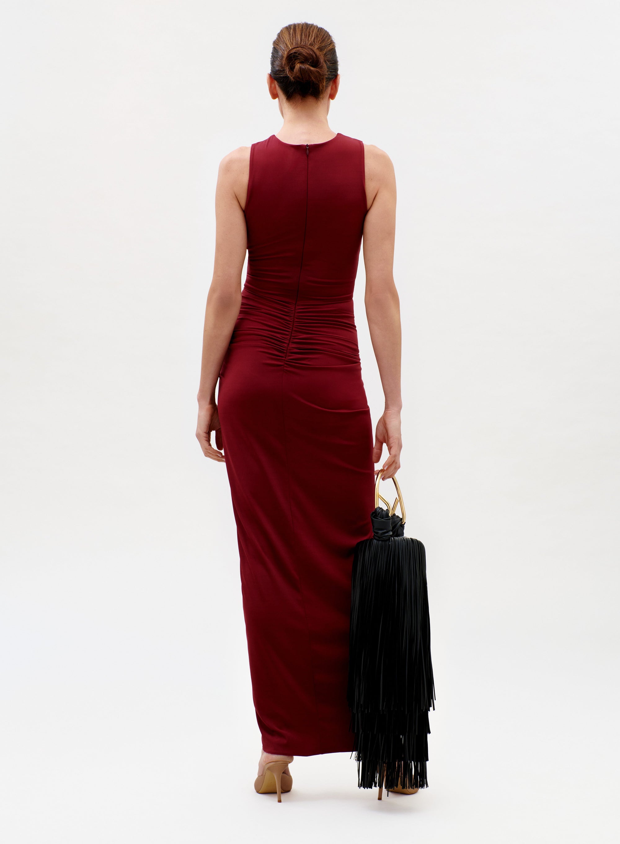 A woman with brown hair in a bun, wearing the sleeveless, form-fitting maroon Leni Dress and beige heels, stands facing away and holds a large black fringed bag with gold handles against a plain white background.