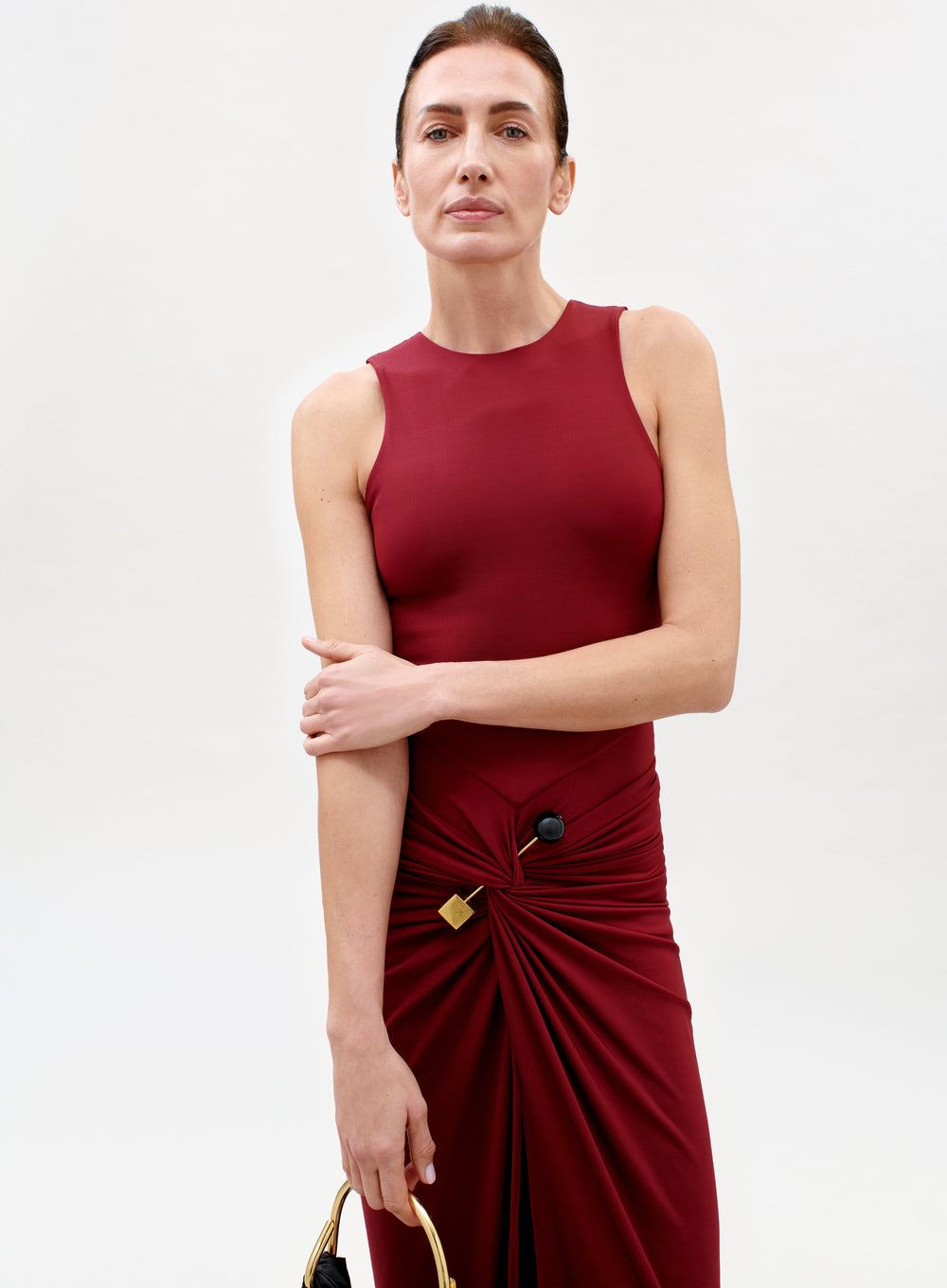 A woman models the sleeveless, fitted Leni Dress in burgundy, featuring gathered fabric and a gold hip accent. She stands against a plain light background, confidently holding a handbag and looking at the camera.
