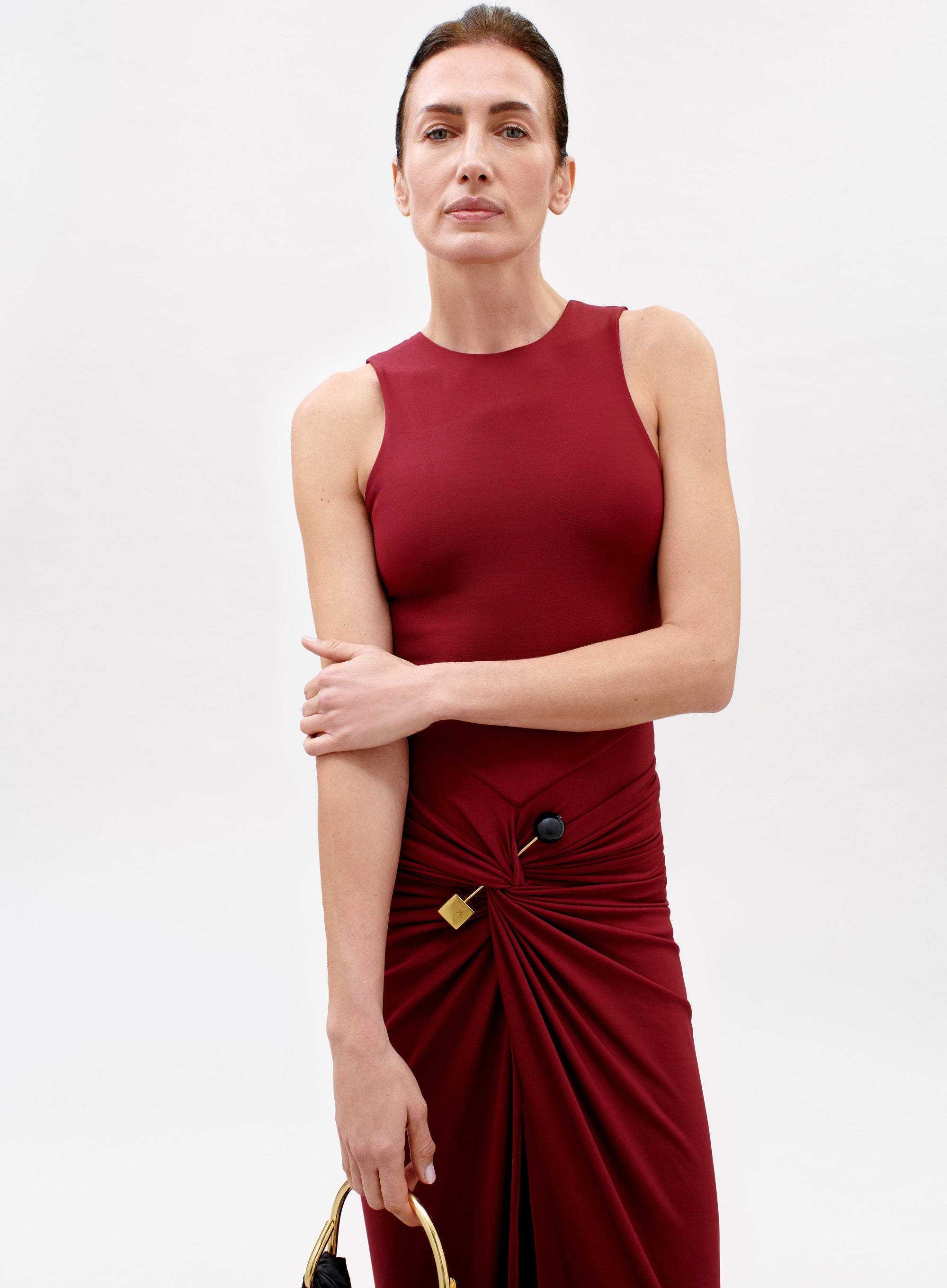 A woman models the sleeveless, fitted Leni Dress in burgundy, featuring gathered fabric and a gold hip accent. She stands against a plain light background, confidently holding a handbag and looking at the camera.