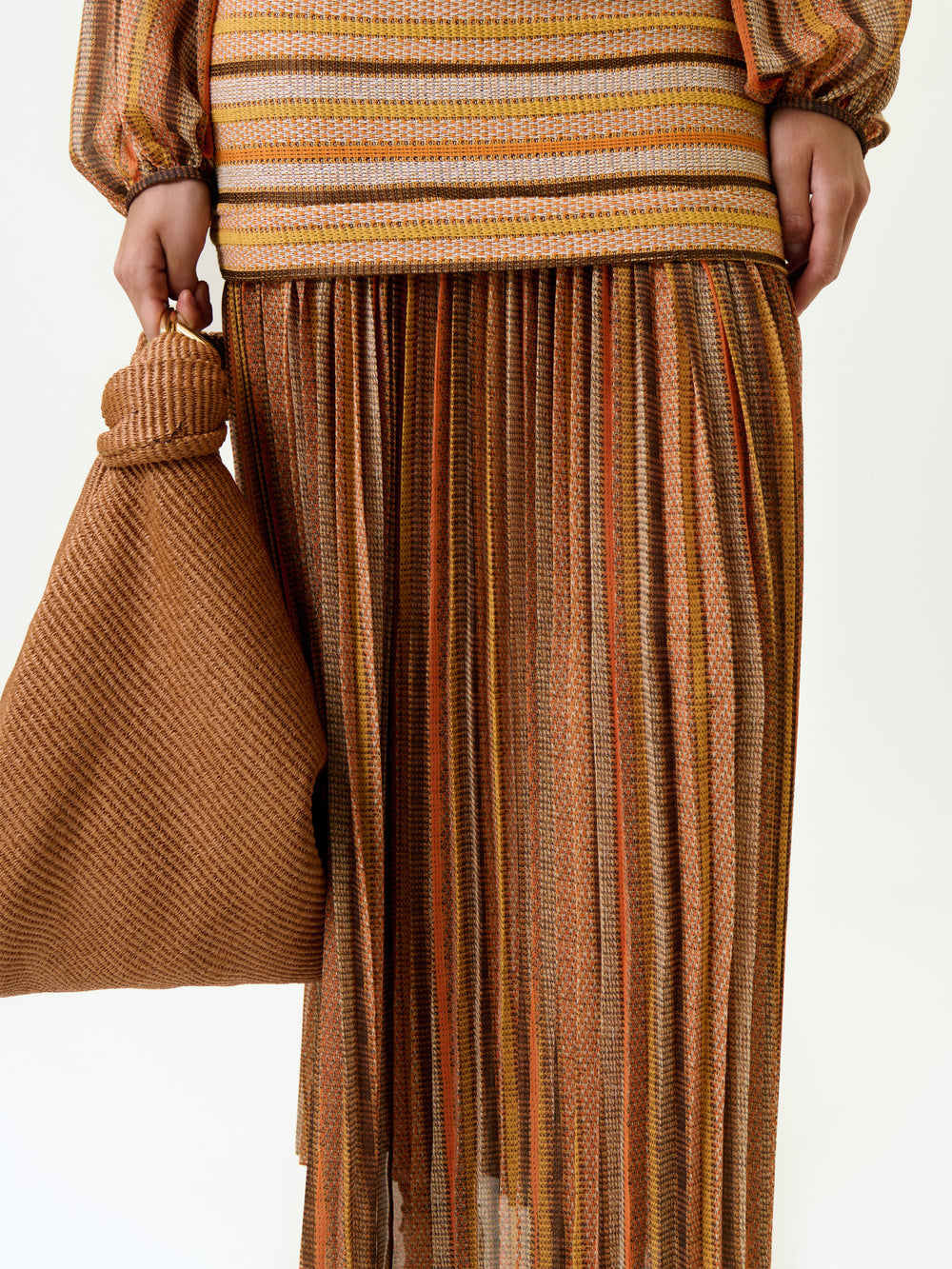 Wearing the Neko Skirt and a long-sleeved top with brown, orange, and yellow stripes, a person holds a textured brown bag. Crafted from Italian fabric for a true-to-size fit, this stylish look is set against a plain background.