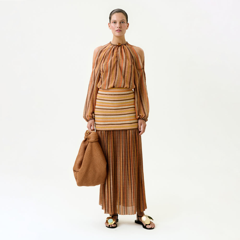 A woman poses against a plain background wearing the Neko Skirt, a striped, pleated maxi in earth tones with a true-to-size fit, paired with a matching cold-shoulder blouse. She carries a large textured brown bag and wears sandals.