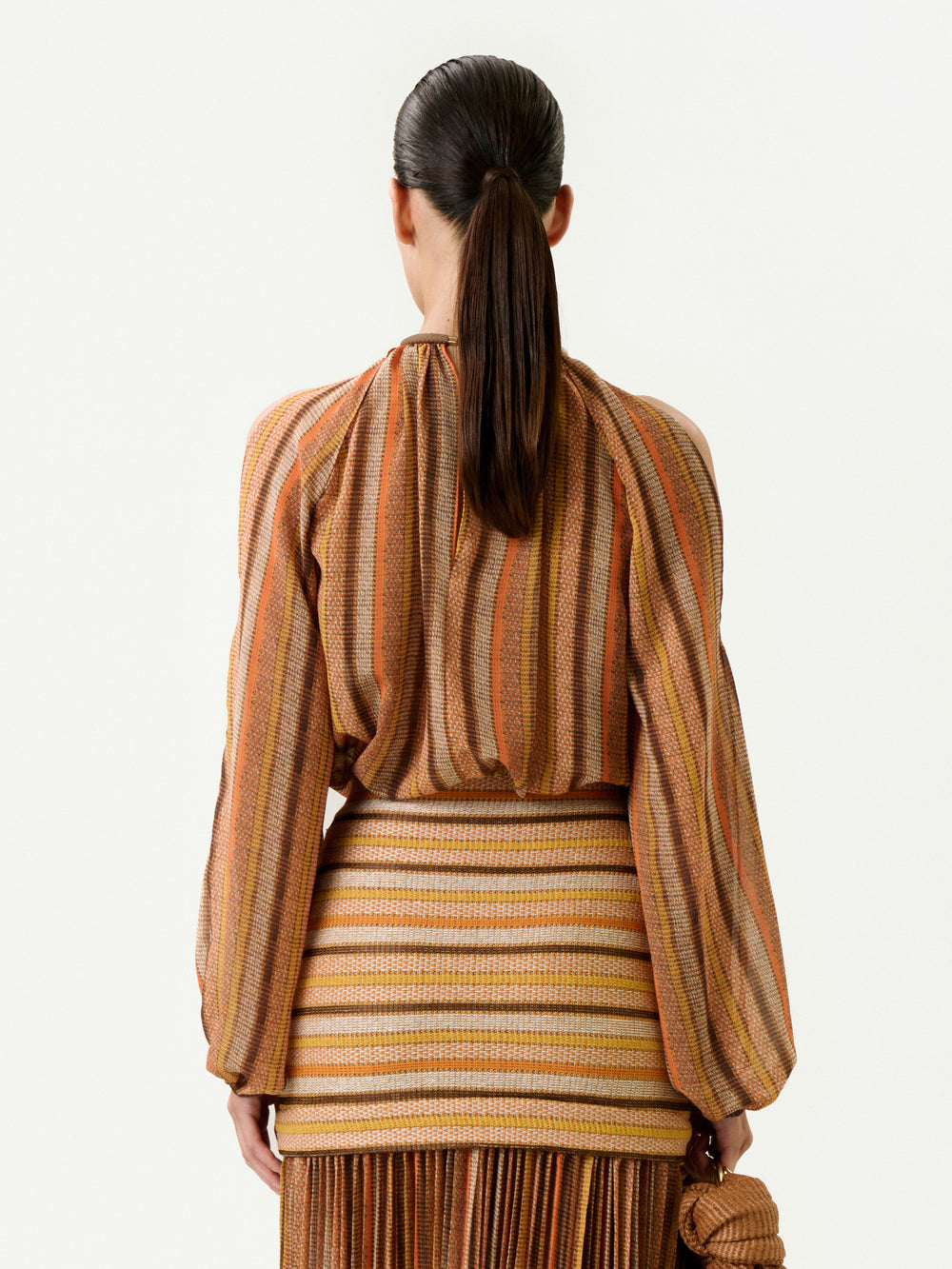 A woman with straight dark hair in a low ponytail is seen from behind, wearing the earth-toned striped Ona Blouse and a fitted skirt made from Italian fabric. She holds a small textured brown bag in her left hand.
