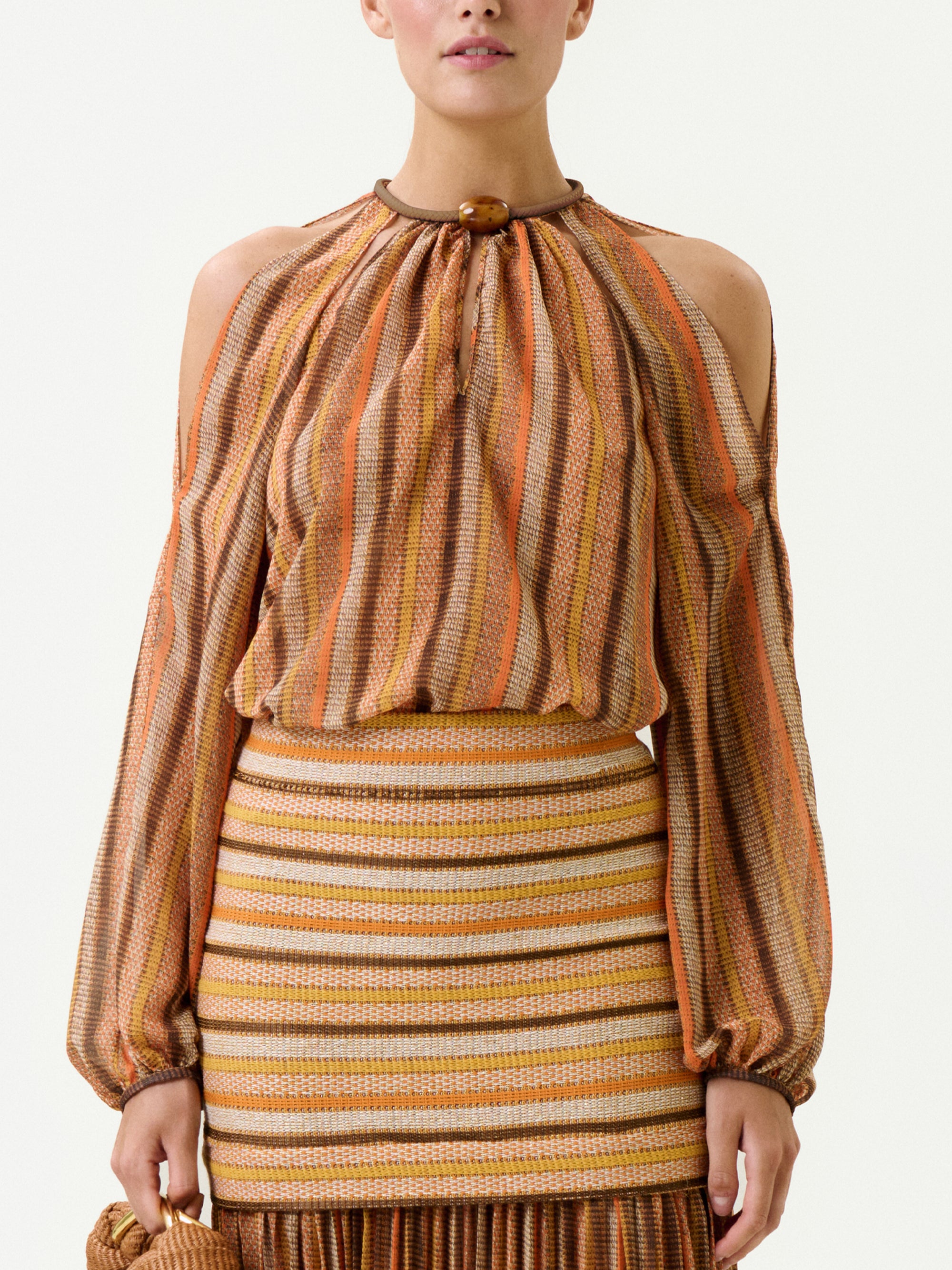 A woman wears the Ona Blouse, a long-sleeved cold-shoulder top with vertical orange, brown, and beige stripes. It’s paired with a matching fitted skirt featuring horizontal stripes and a fringed hem. Made from 100% Italian polyester fabric. She carries a small bag.