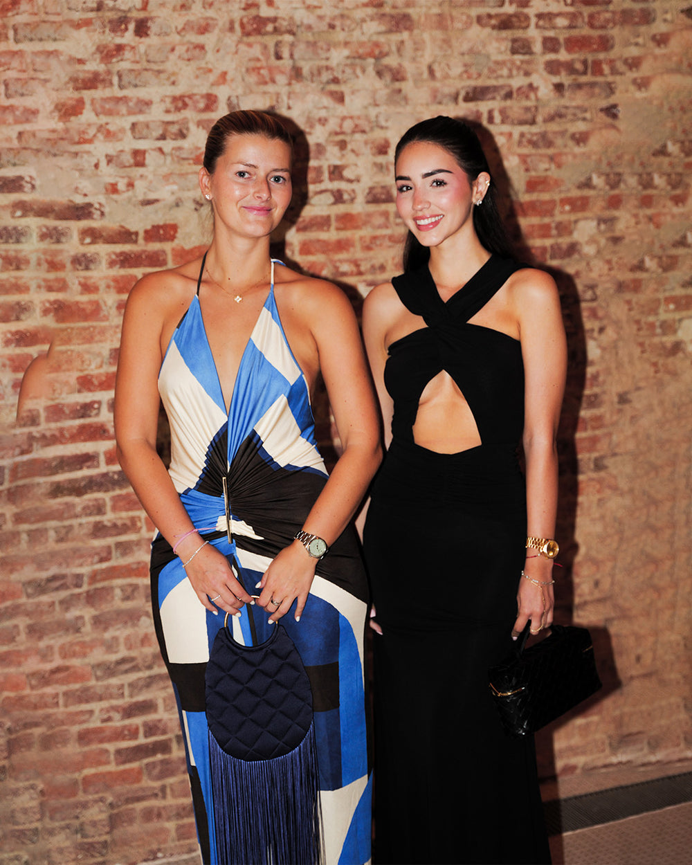 Two women posing together in front of a brick wall. The woman on the left wears a blue, white, and black geometric halter dress and holds a black quilted clutch.