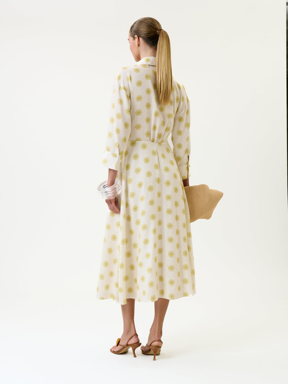 Woman with long ponytail stands facing away on a plain white background, wearing the Milene Blouse, paired with tan heels and holding a woven clutch.