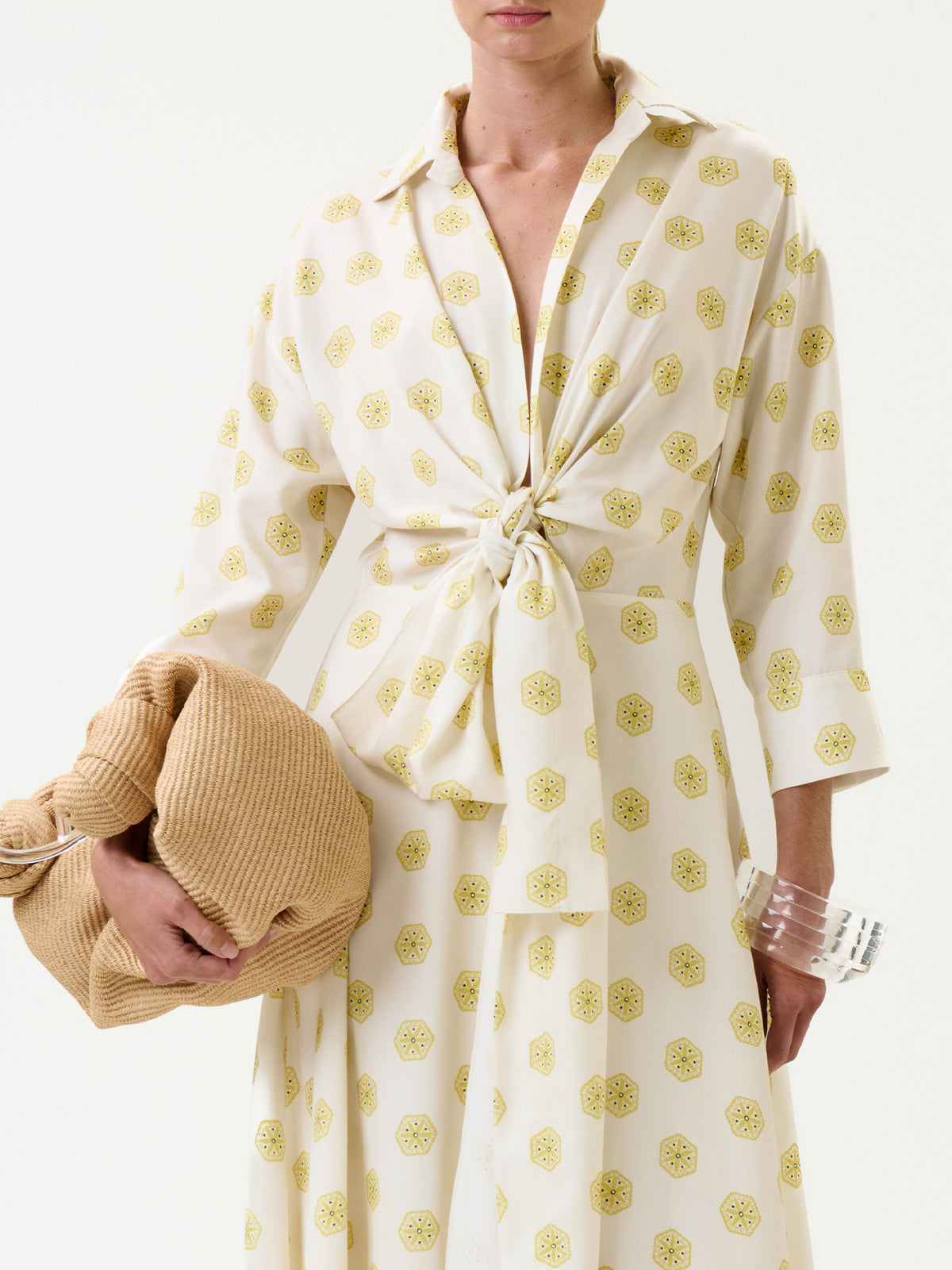 A woman wears the Milene Blouse, a cream top with yellow patterns and a front tie, paired with a chunky clear bracelet and holding a beige textured item. The background is plain white.