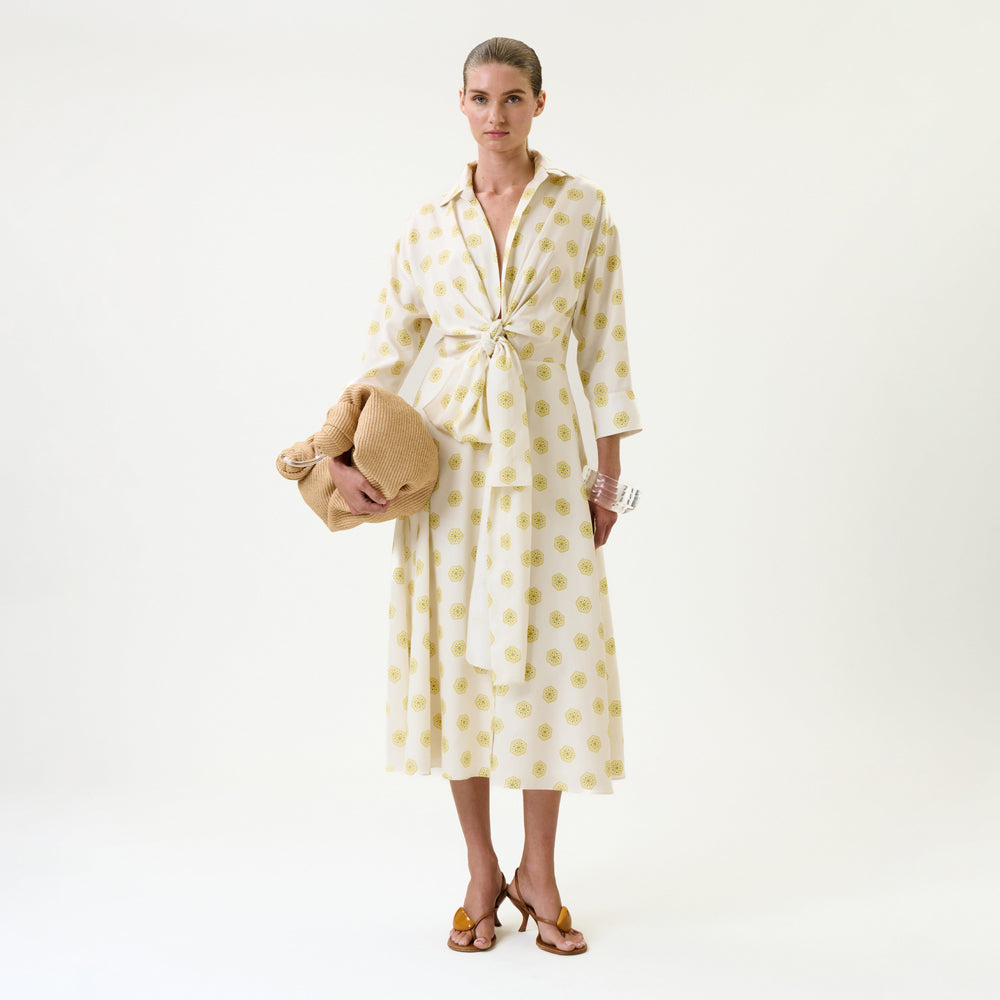 A woman stands against a plain white background wearing the Milene Blouse, styled with heeled sandals and her hair pulled back. She holds a tan blanket or bag.