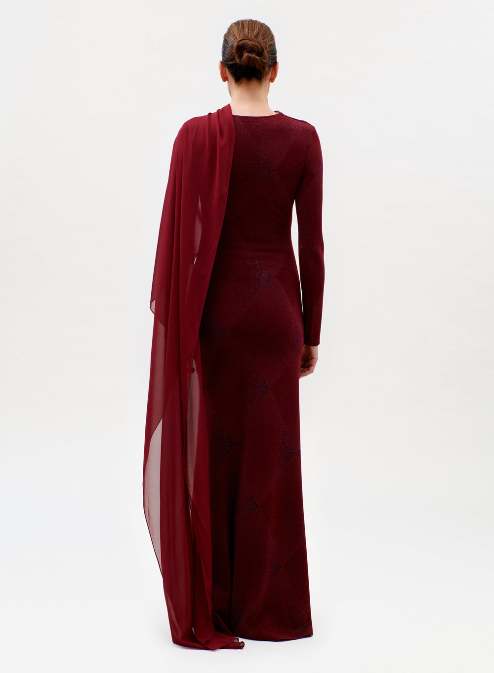 A woman, her hair in a neat bun, faces away while wearing the floor-length, long-sleeved Moon Dress in dark red with a sheer matching drape over one shoulder, set against a plain white background.