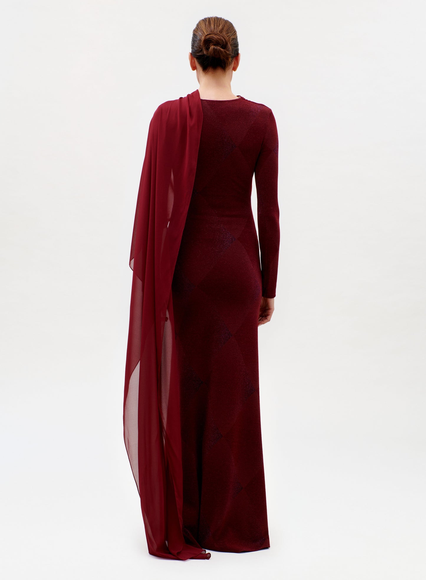 A woman, her hair in a neat bun, faces away while wearing the floor-length, long-sleeved Moon Dress in dark red with a sheer matching drape over one shoulder, set against a plain white background.