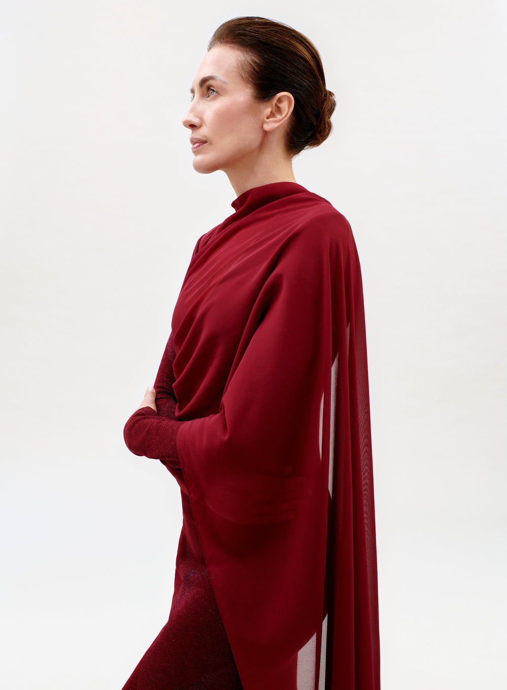 A woman with brown hair in a bun, wearing the elegant red Moon Dress with a matching shawl, stands in profile against a plain white background.