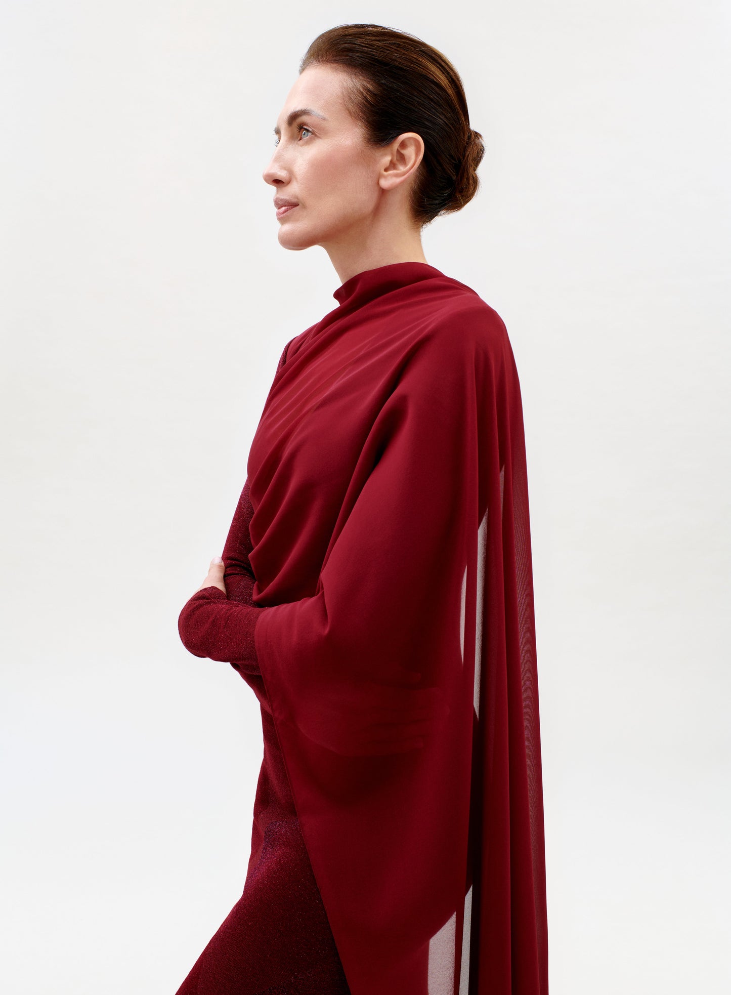 A woman with brown hair in a bun, wearing the elegant red Moon Dress with a matching shawl, stands in profile against a plain white background.