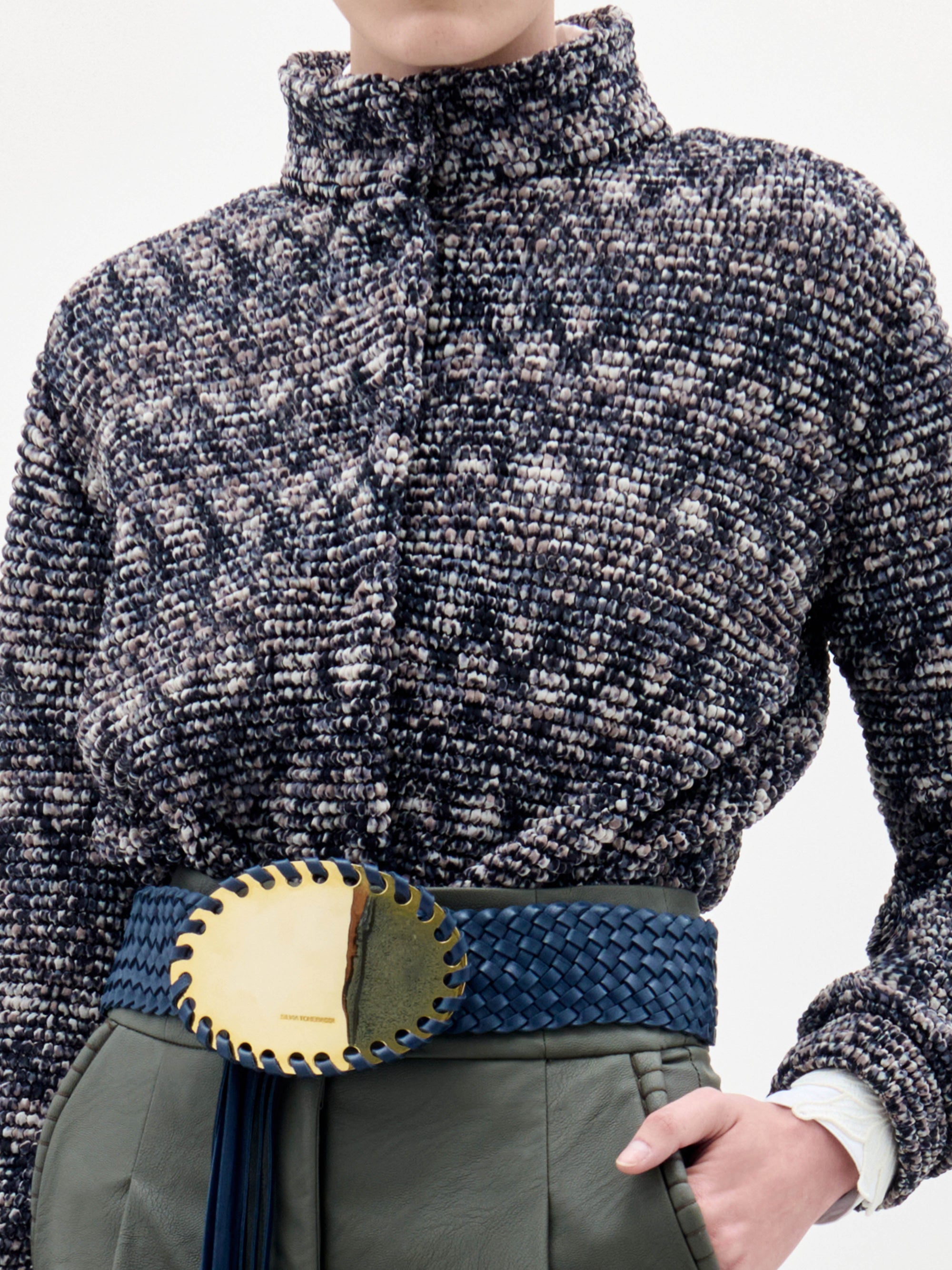 A person stands against a plain background wearing the Zalta Jacket, olive green pants, and a wide blue belt with a large gold buckle.