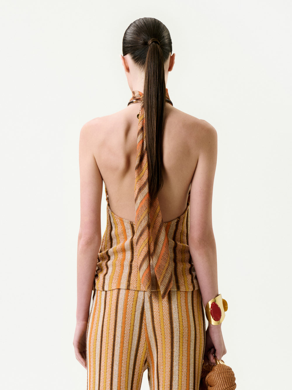 A person with long, dark hair in a ponytail is seen from behind wearing the Mitzy Blouse—a backless, striped halter top in warm tones made from Italian fabric—styled with matching pants, a chunky bracelet, and a textured bag.