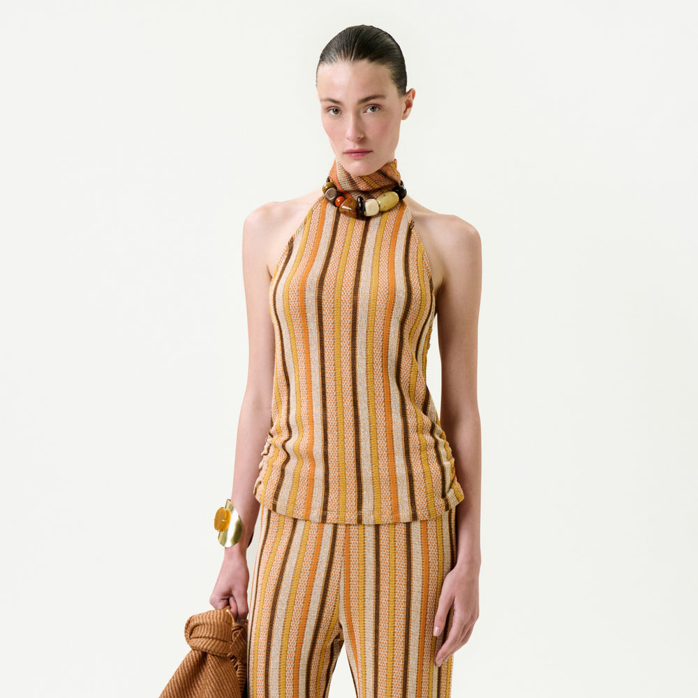 A woman stands against a plain background wearing the Mitzy Blouse, a sleeveless high-neck top featuring vertical yellow, beige, and orange stripes. Made from Italian fabric, it offers a true-to-size fit. She pairs it with matching pants and accessorizes with a chunky bracelet.