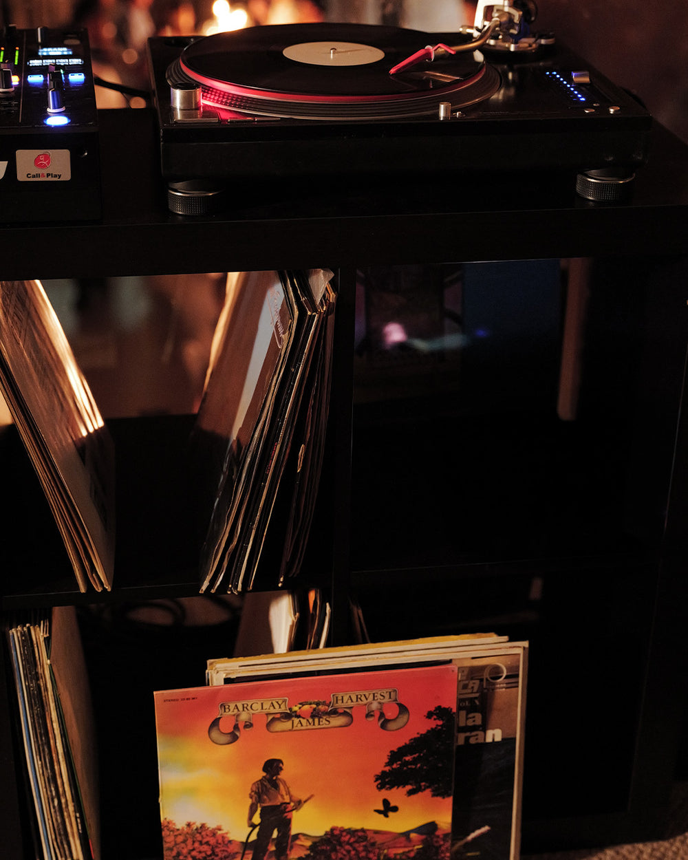 A black turntable with a vinyl record playing sits on a dark shelf in a dimly lit room, with vinyl records stacked in the background and a colorful album cover visible in the foreground.