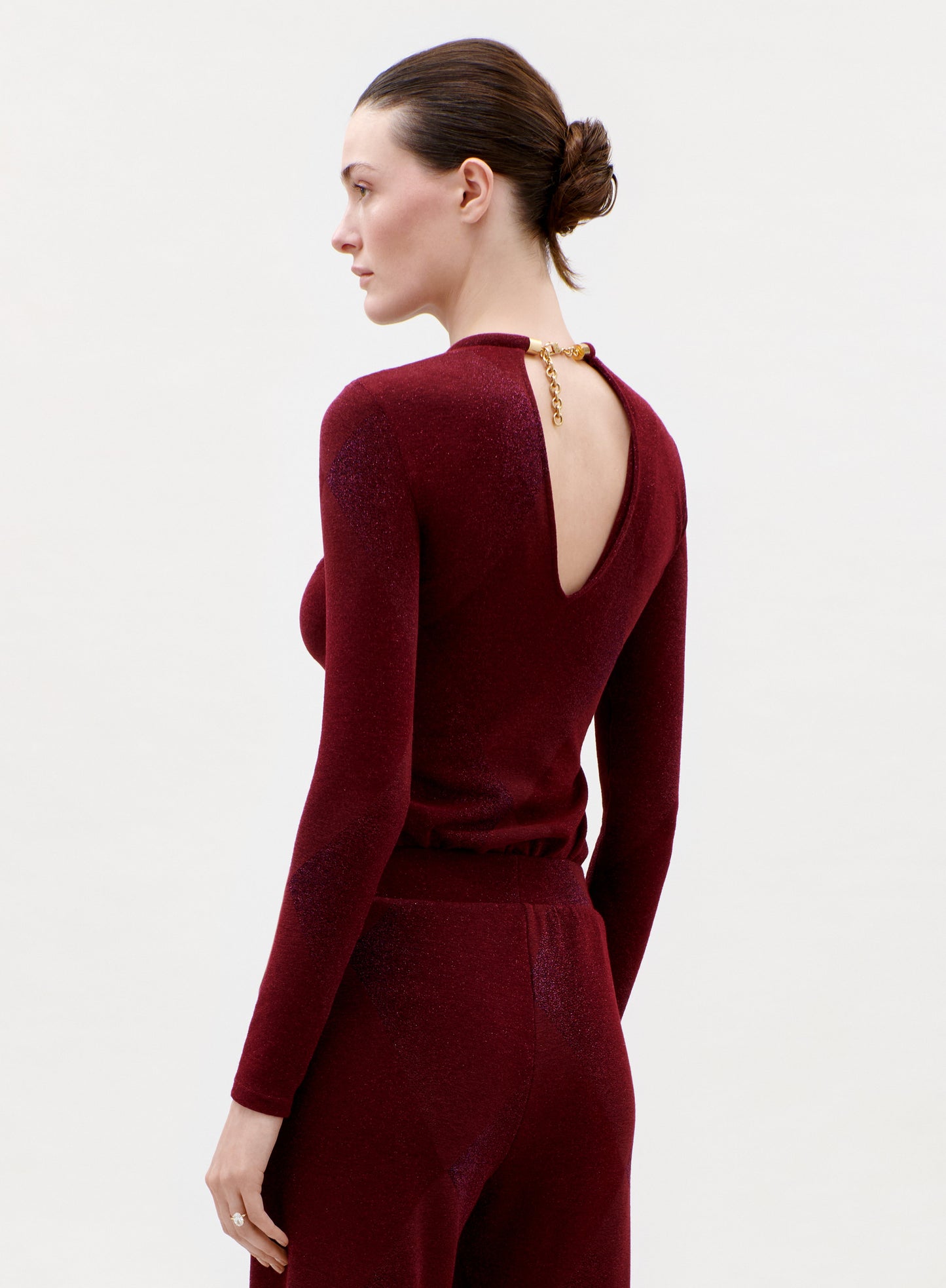 A woman with dark hair in a low bun wears the fitted, long-sleeved Lalia T-Shirt in burgundy with an open back and gold clasp, paired with matching pants, posing against a plain light background.
