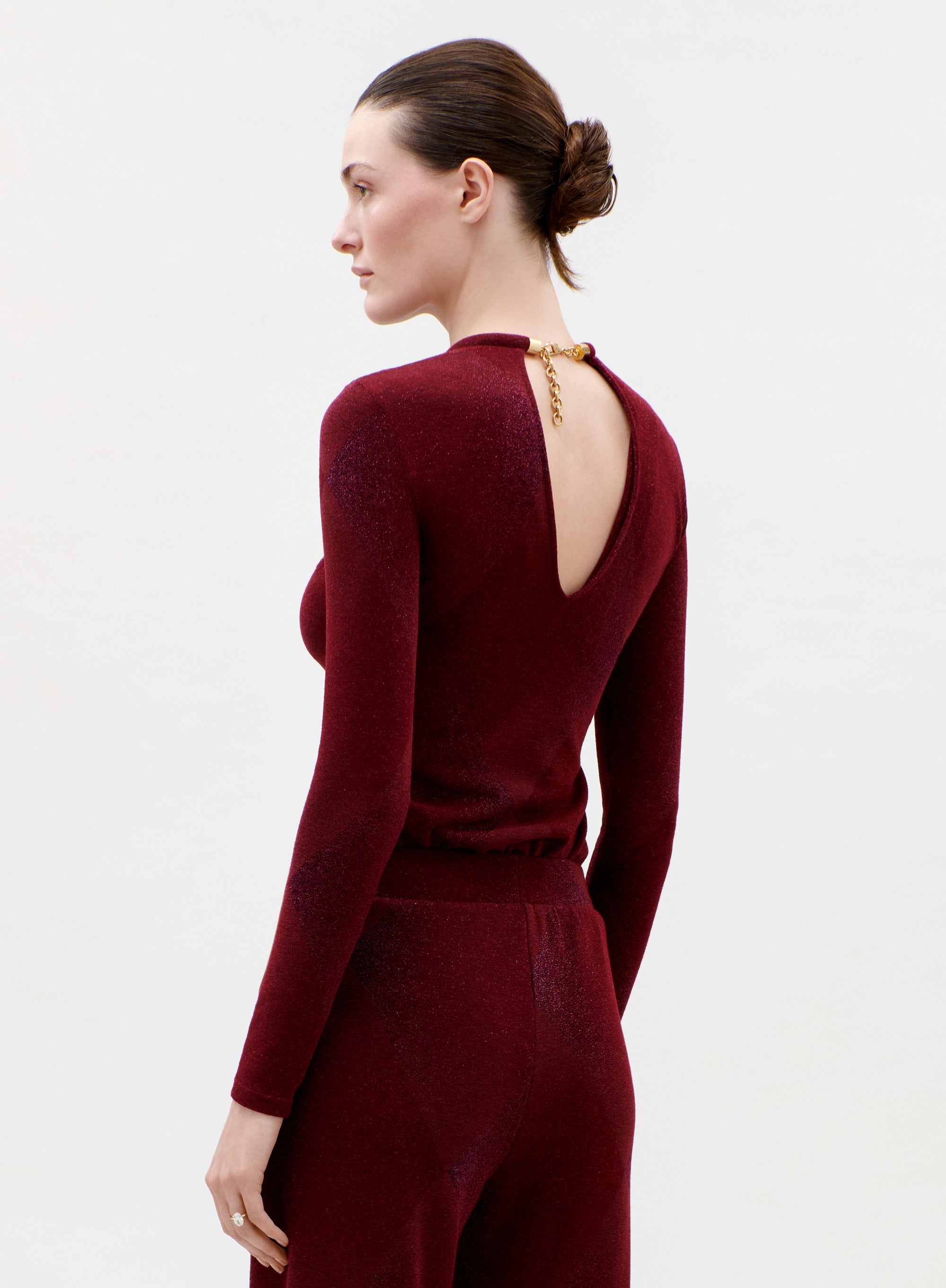 A woman with dark hair in a low bun wears the fitted, long-sleeved Lalia T-Shirt in burgundy with an open back and gold clasp, paired with matching pants, posing against a plain light background.