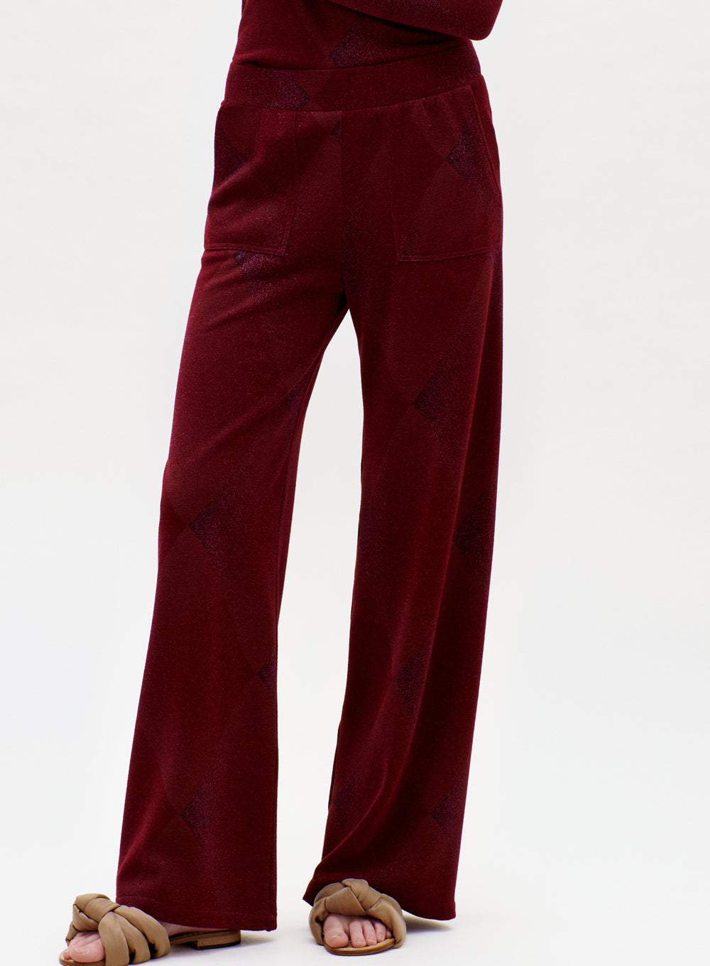 The lower half of a person is shown wearing the Colbie Pant in a loose-fitting, deep red style with pockets, paired with brown knotted sandals against a plain background.