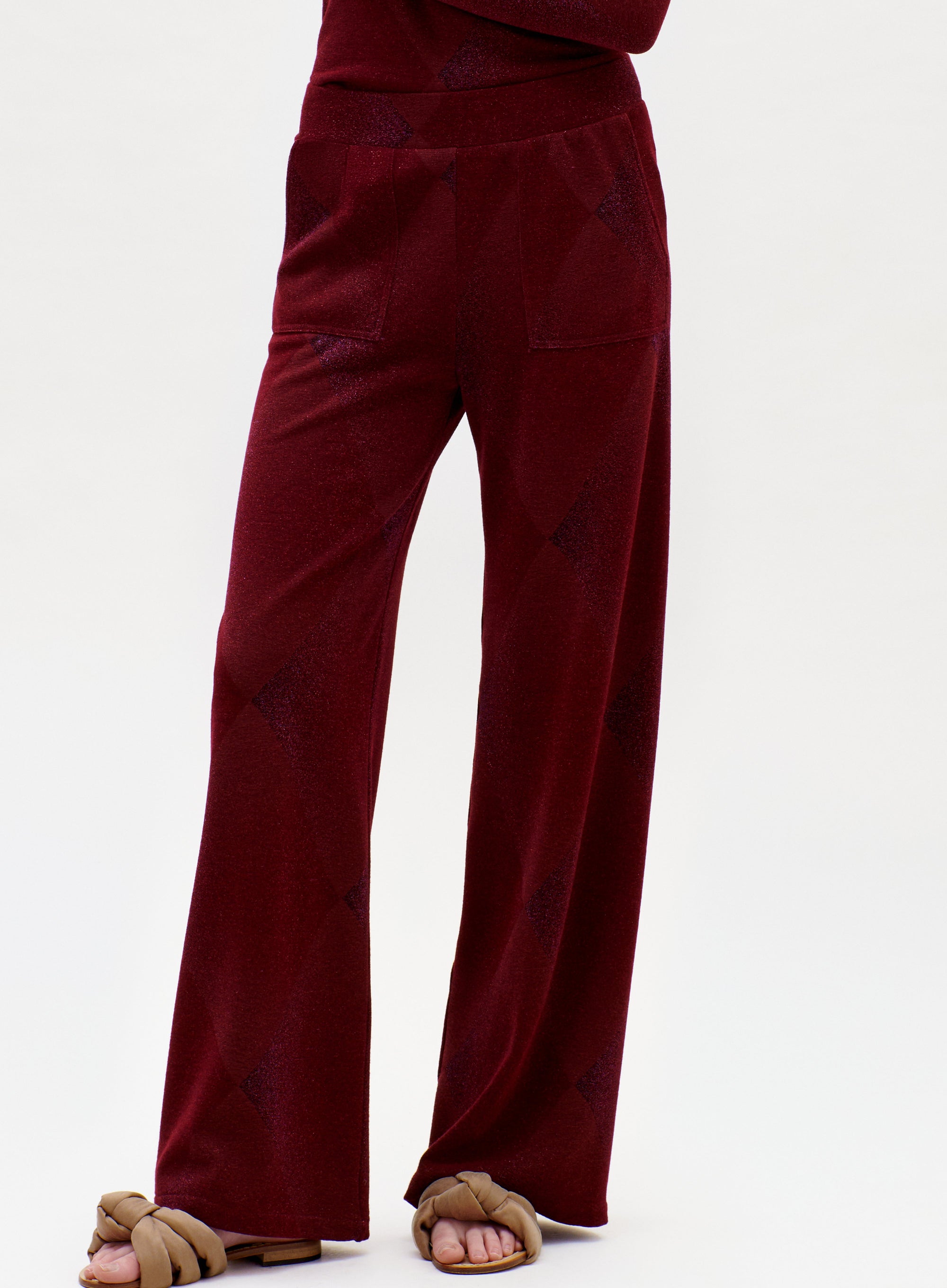 The lower half of a person is shown wearing the Colbie Pant in a loose-fitting, deep red style with pockets, paired with brown knotted sandals against a plain background.