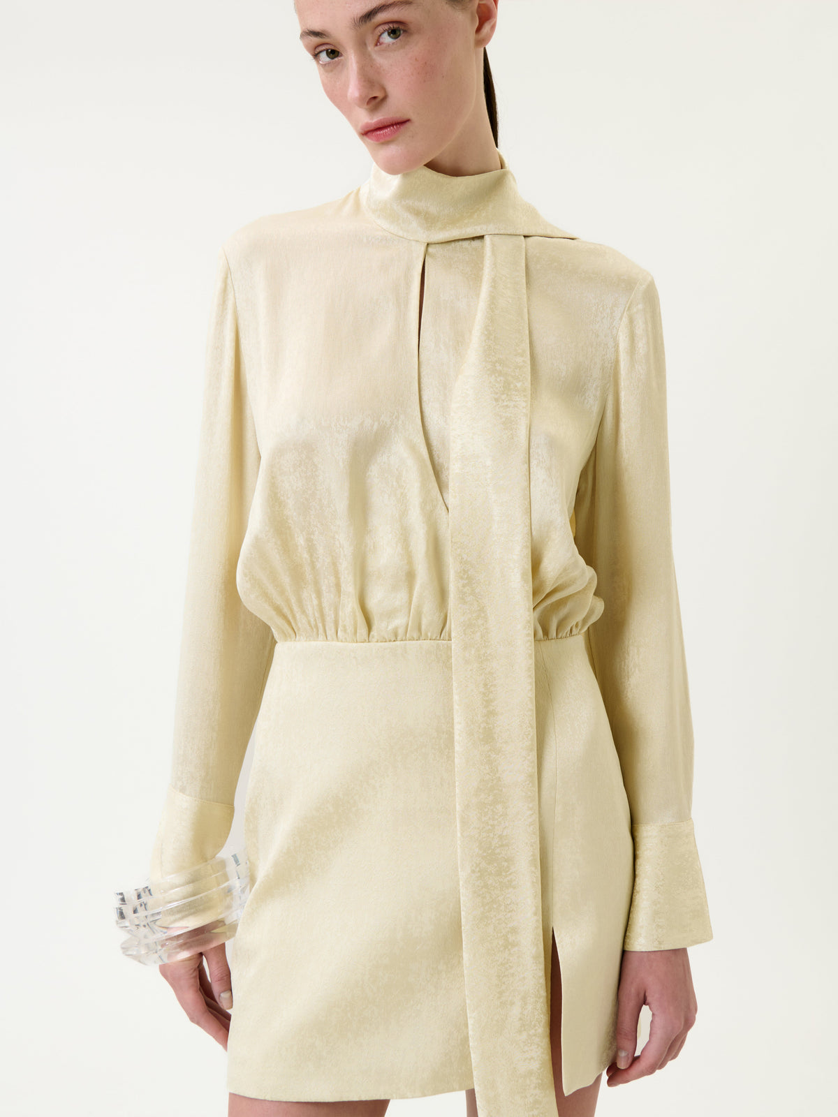 A person is wearing the Imelda Dress, a light yellow satin piece with long sleeves, a high neck, and a loose tie detail. The dress features a fitted waist and a side slit. They are standing against a plain white background and accessorizing with clear, chunky bracelets.
