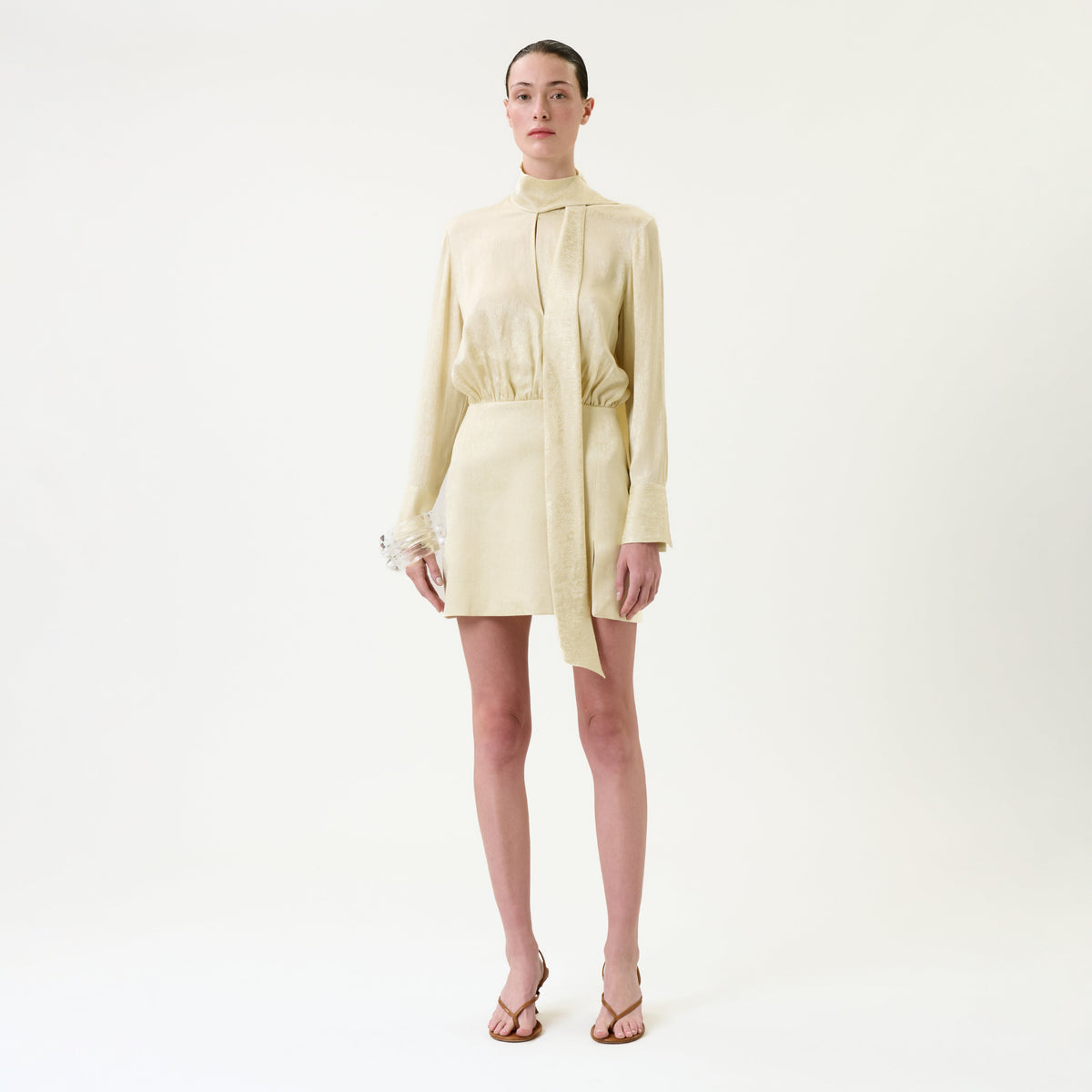 A person stands against a plain white background, wearing the Imelda Dress— a long-sleeved, light beige mini dress with a high collar and tie detail—paired with tan sandals.