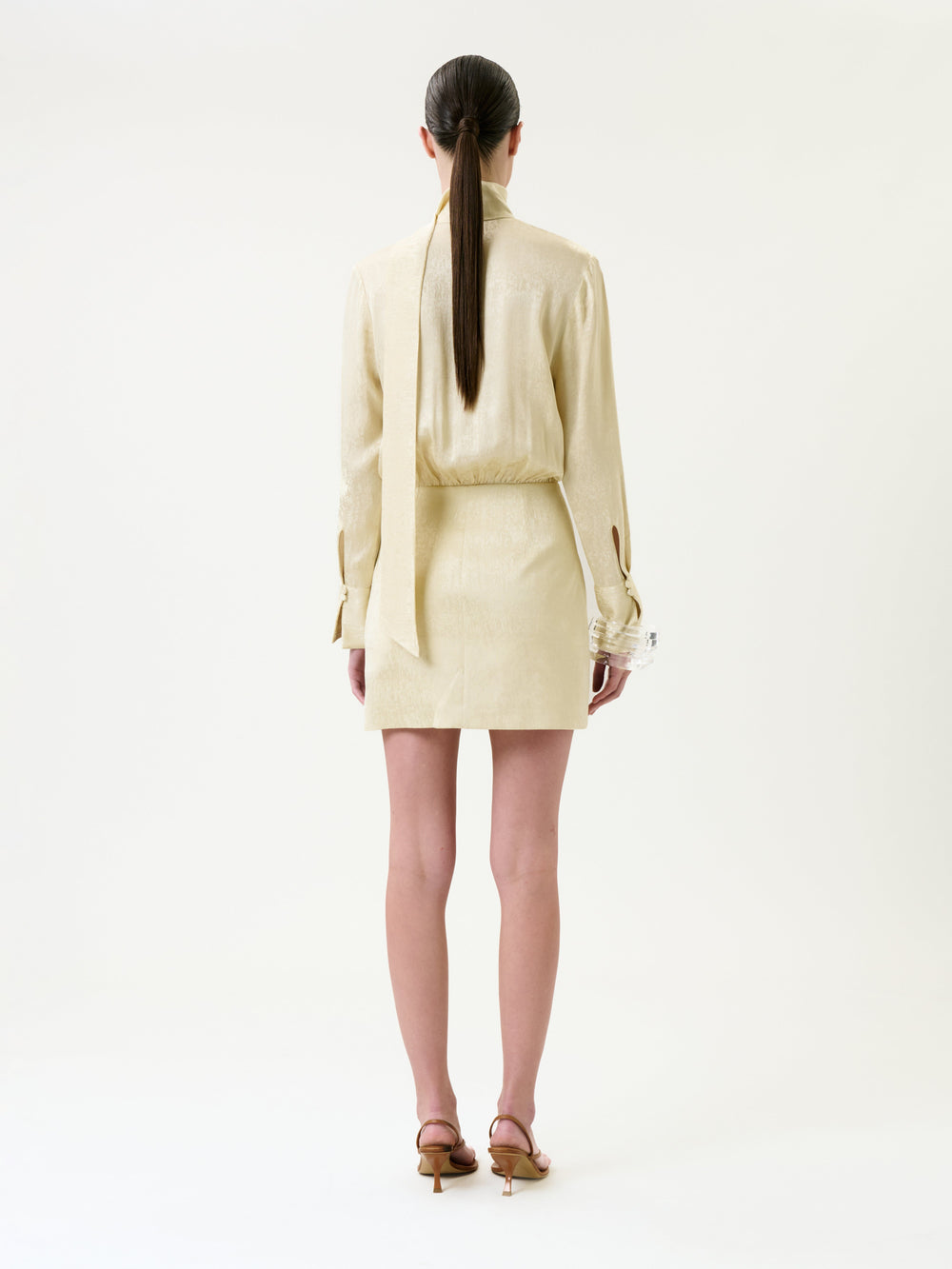 A woman stands facing away, wearing the Imelda Dress in light beige with long sleeves and a tied scarf detail, paired with heeled sandals. Her straight, dark hair is styled in a low ponytail.