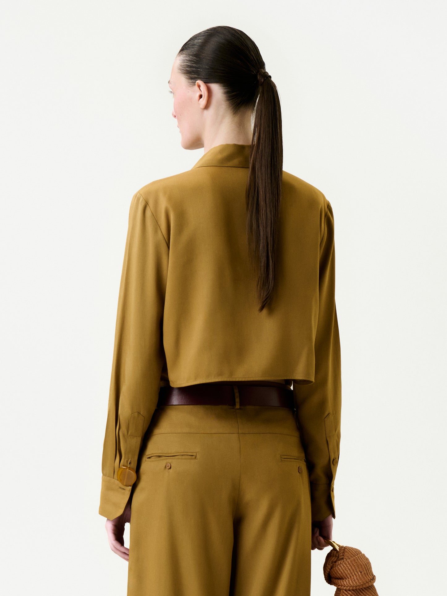A person with long brown hair in a ponytail is shown from behind, wearing the mustard-colored Mazz Blouse with matching trousers and holding a small brown textured clutch bag in their right hand.